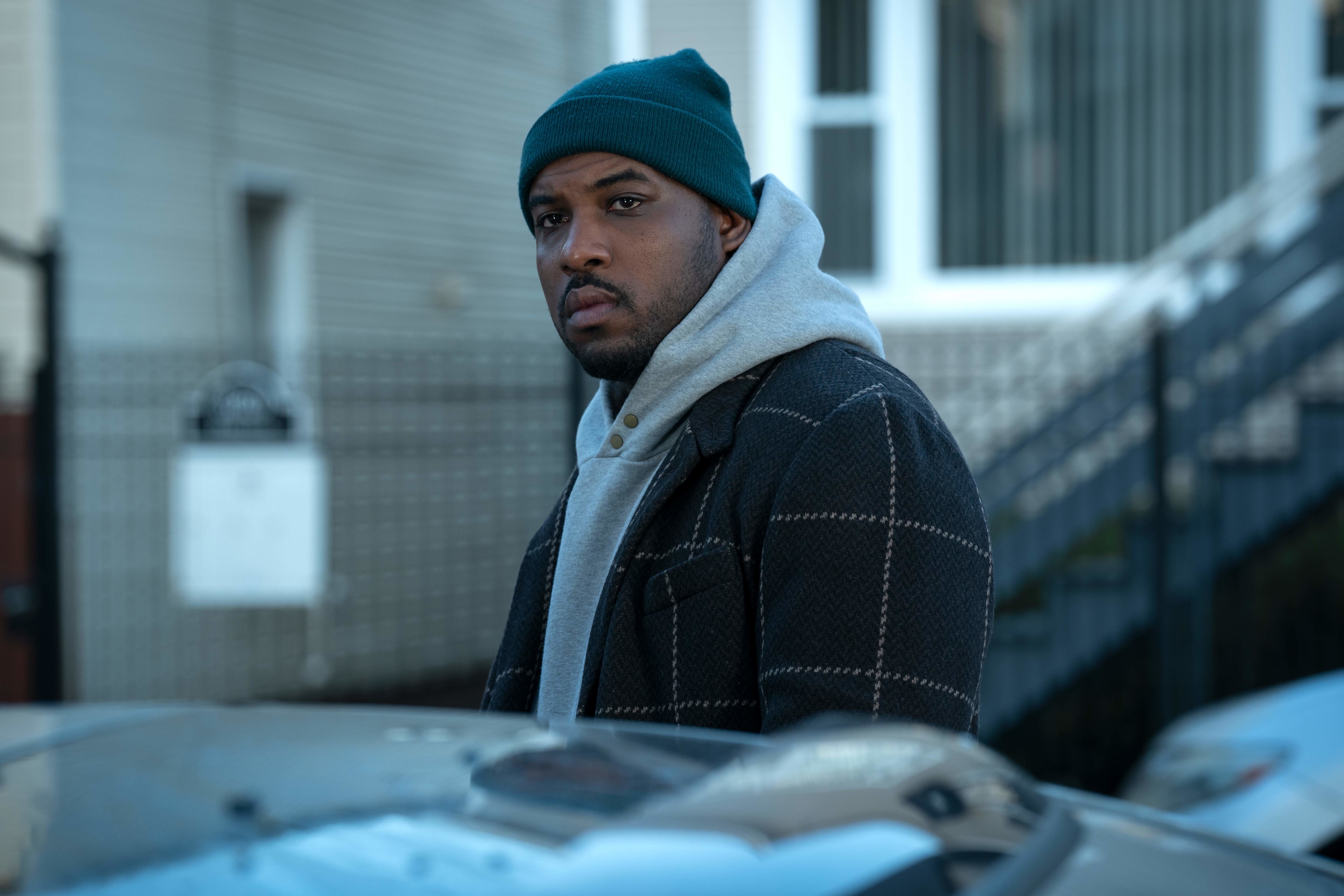Lionel Boyce wearing a beanie, dark coat, and hoodie in The Bear as she stands by a car
