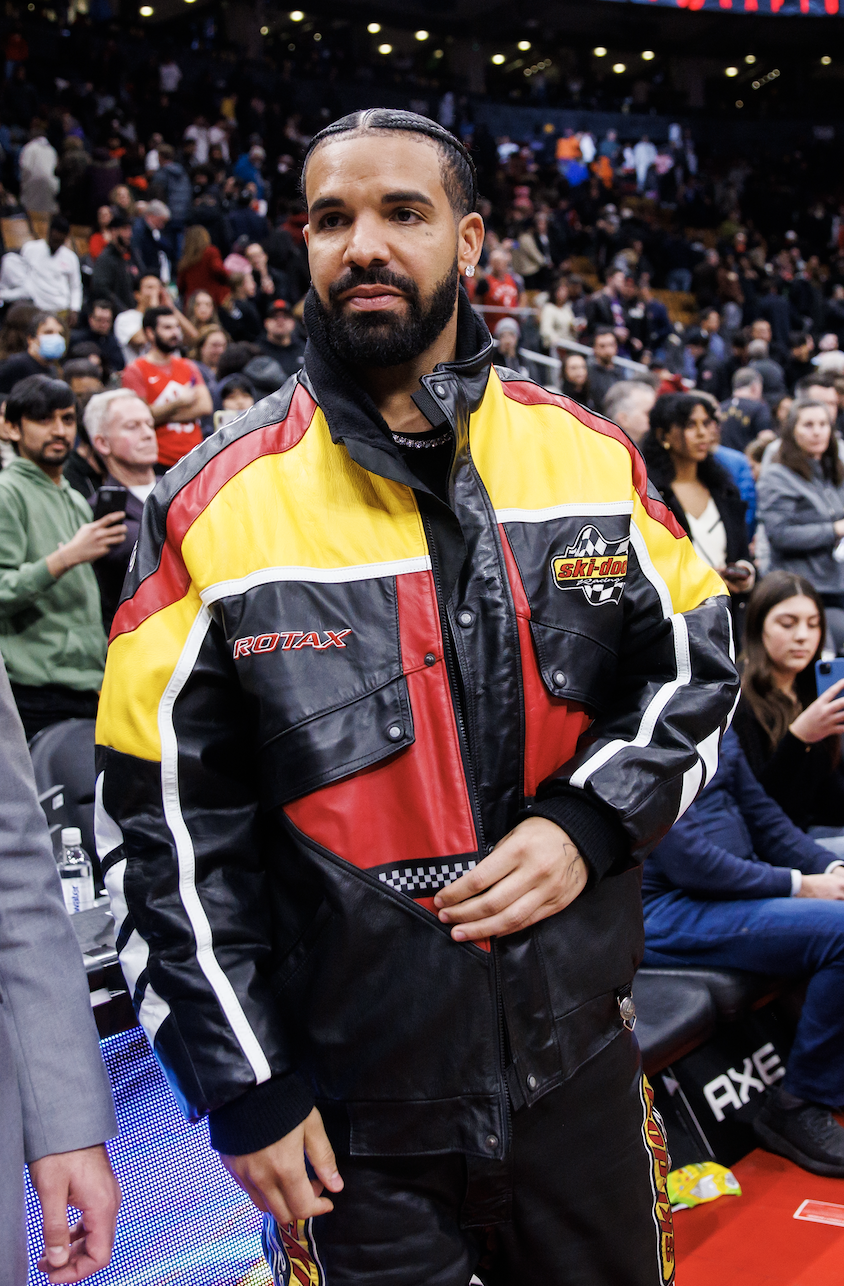 Drake standing court-side at a basketball game wearing a motorcycle jacket