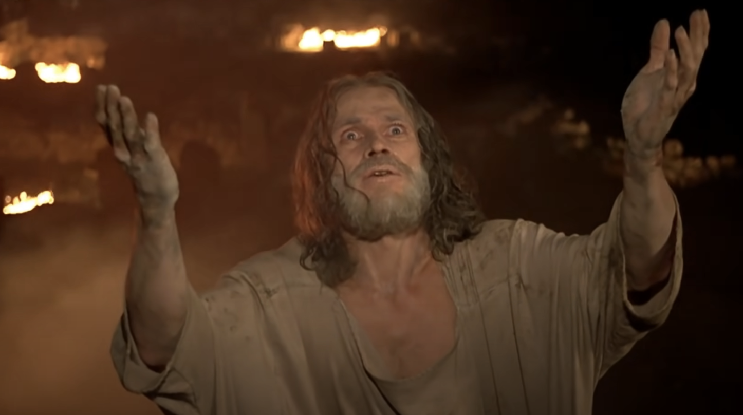 A man with long hair and a beard, wearing tattered clothing, stands with arms raised and a look of anguish on his face, with fire visible in the background