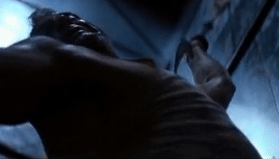 A person, dressed in a sleeveless shirt, hangs from an overhead support, swinging using their arms in an intense, cinematic action scene