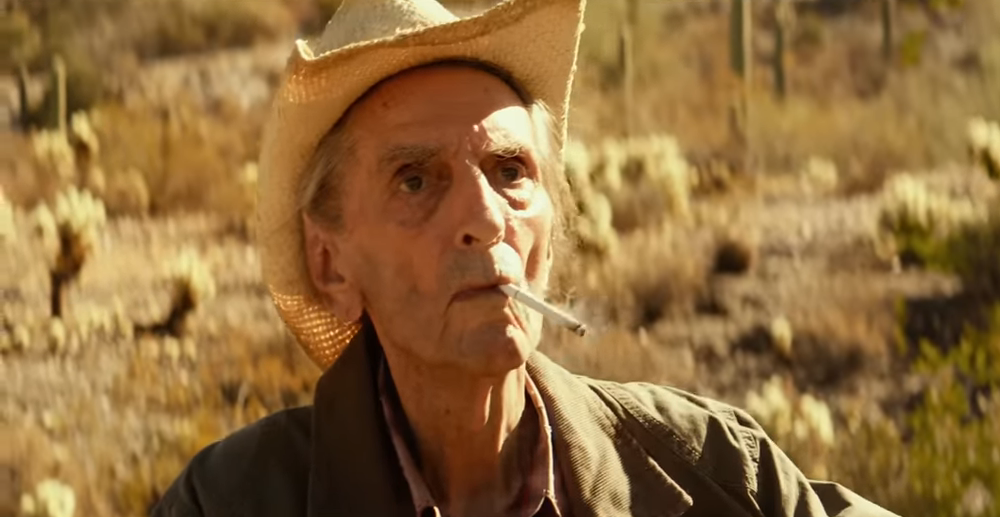 Stanton in worn out clothes and a cowboy hat smoking a cigarette in lucky