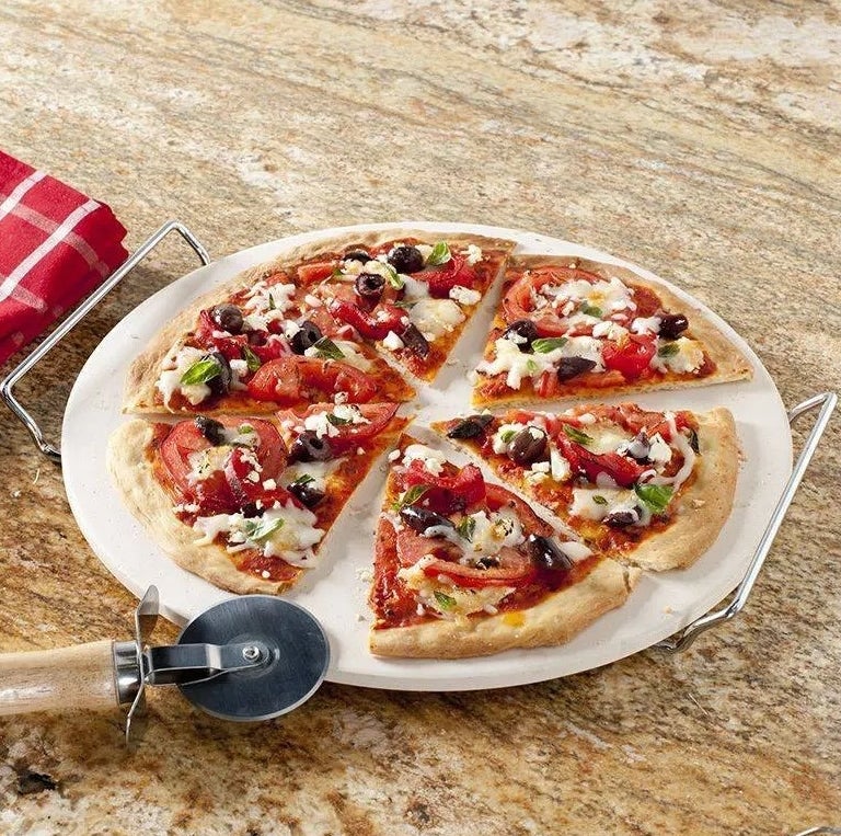 The included pizza cutter next to the pizza stand and stone topped with a cooked pizza.