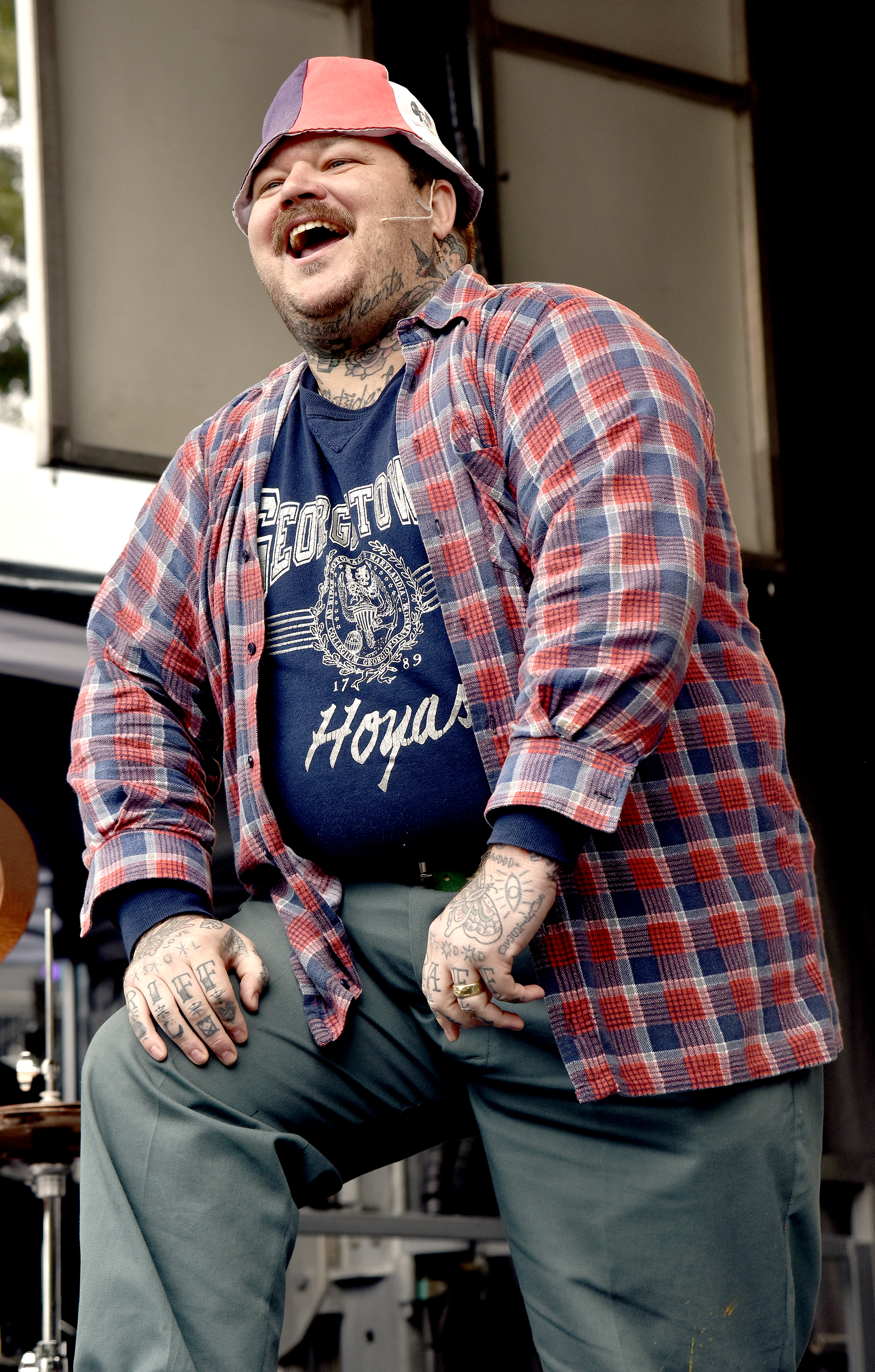 A heavily tattooed Matty Matheson at a cooking demonstration wearing a Georgetown Hoyas t-shirt, a plaid shirt over that, and pants. He&#x27;s also wearing a bucket hat
