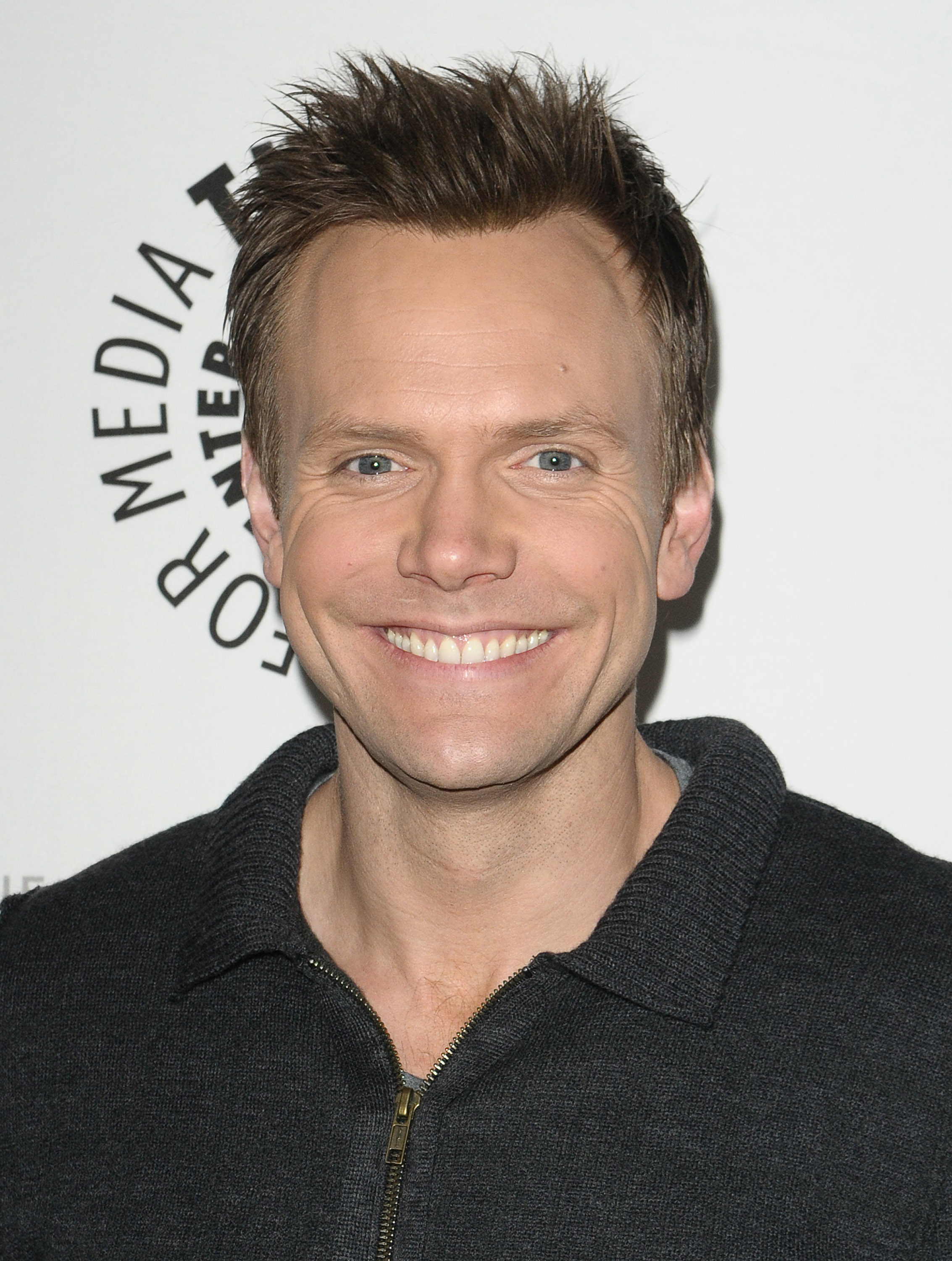 A closeup of Joel McHale smiling on the red carpet of a media event. He&#x27;s wearing a zippered sweater