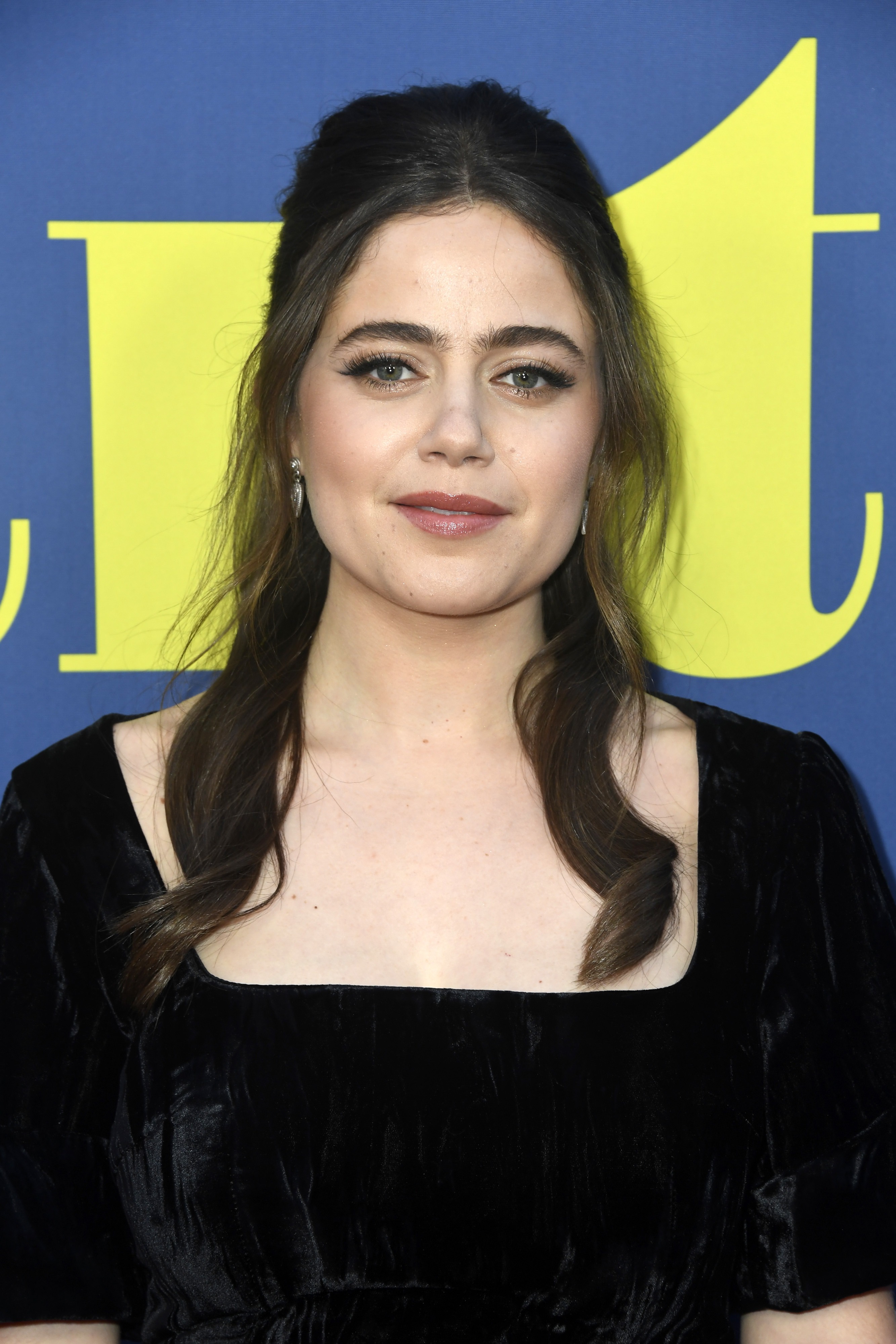 A closeup of Molly Gordon at a screening. Her brunette hair is past her shoulders and styled half up, half down