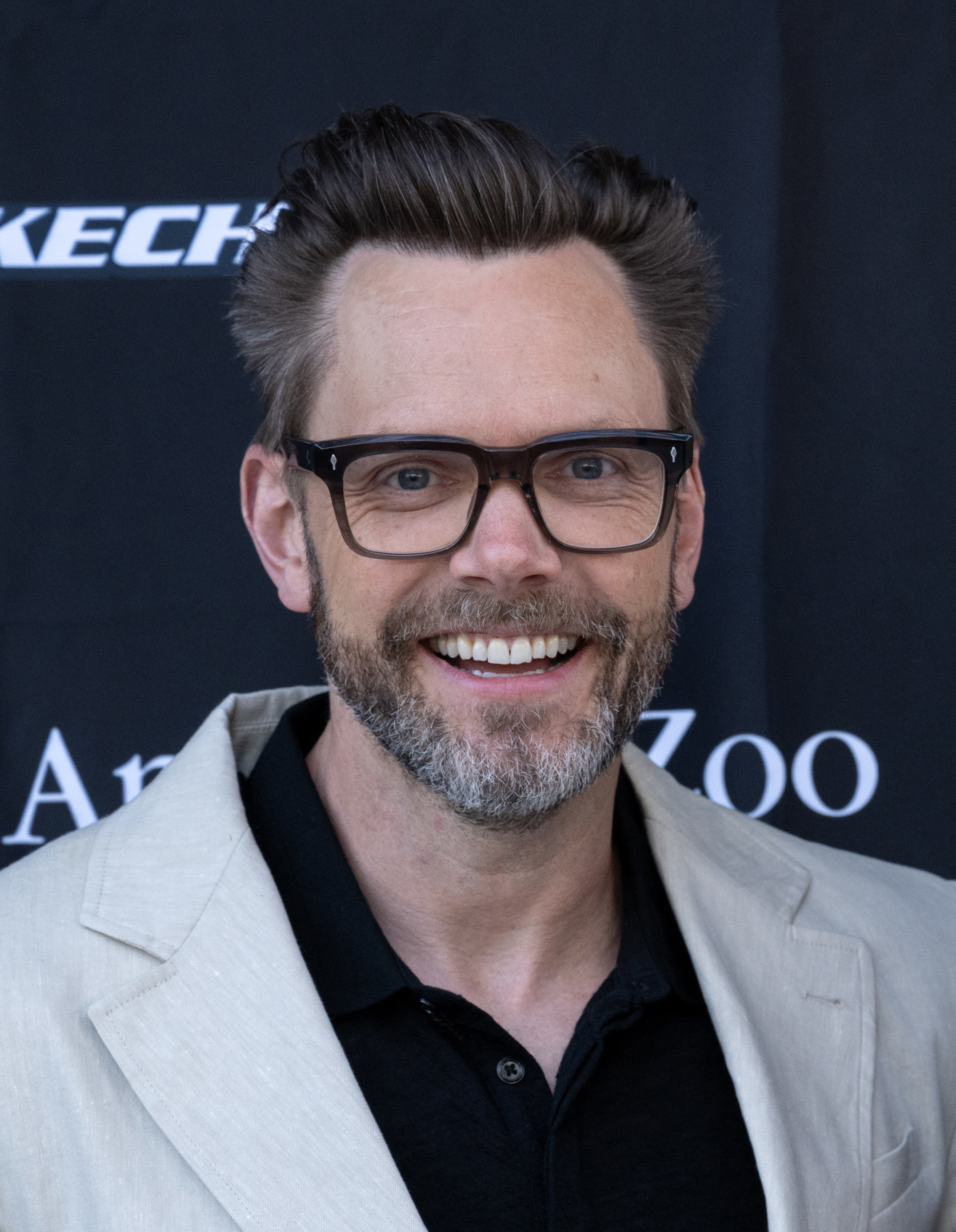 A closeup of Joel McHale smiling at a media event. He&#x27;s wearing a casual suit and eyeglasses