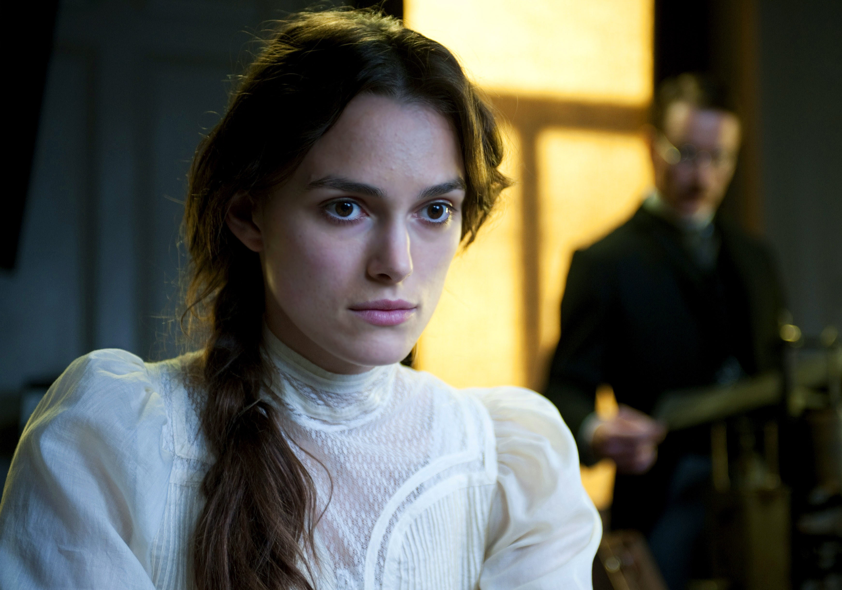 Keira Knightley stares off into space while a rigid man stands far behind her in a dark room