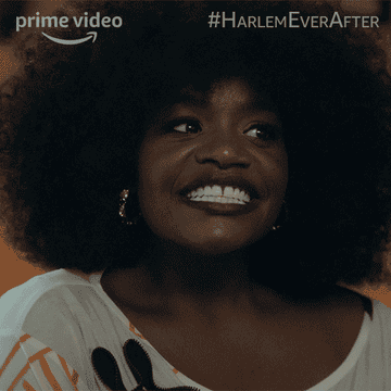 A Black woman with a beautiful big afro, smiling and then immediately recoiling as if something uncomfortable was said.