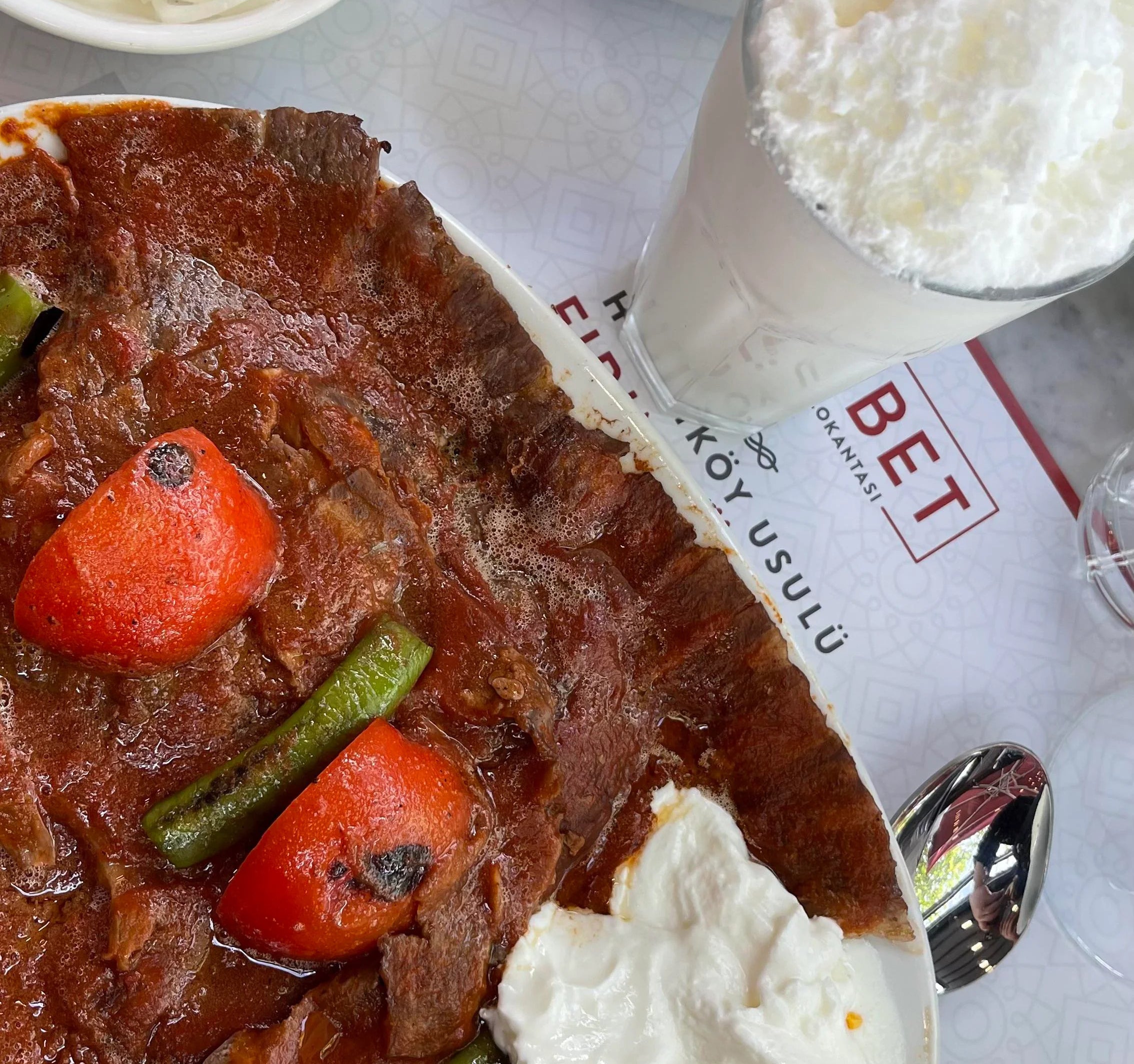 A cup of ayran is on the table next to a plate of doner iskender and pickled veggies