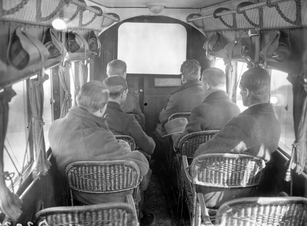 Men sitting in wicker-type chairs facing a screen, with a very, very narrow aisle between the two rows
