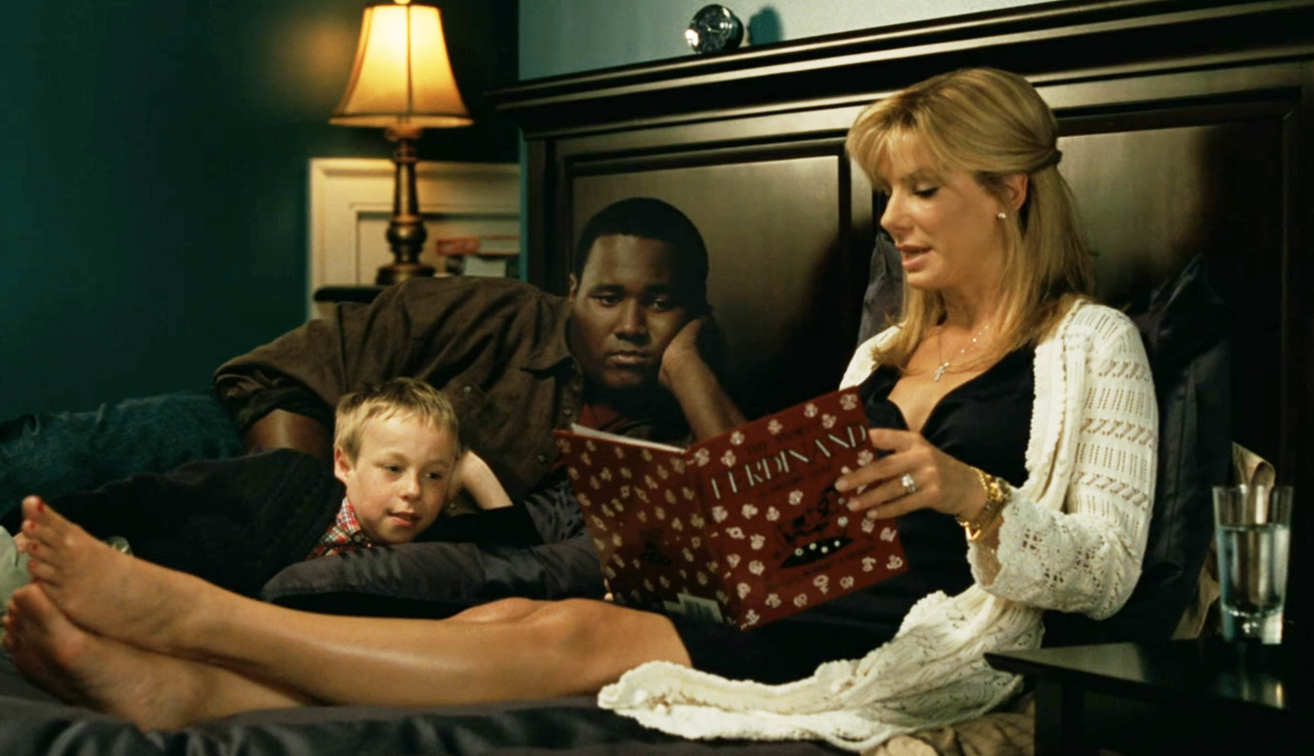 Michael Oher laying in bed as Leigh Anne Tuohy reads to him and her son in a scene from the film