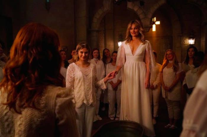 Alice and Polly, two blonde women wearing all white, hold hands in the centre of a room. Multiple people, also dressed in white, surround them.
