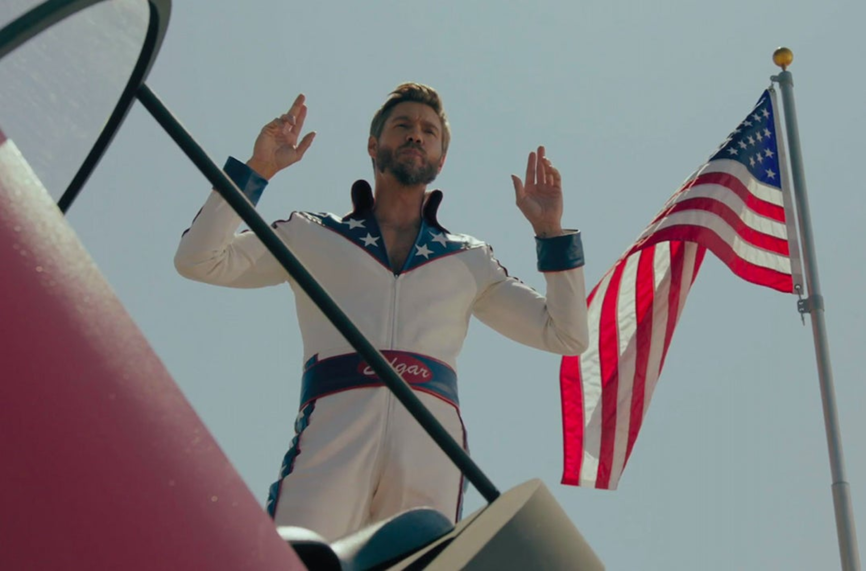 Edgar Evernever, a man with short dirty blonde hair and a beard, stands with his hands in the air. He is wearing a white bodysuit, with blue and red details. The flag of the USA stands next to him.
