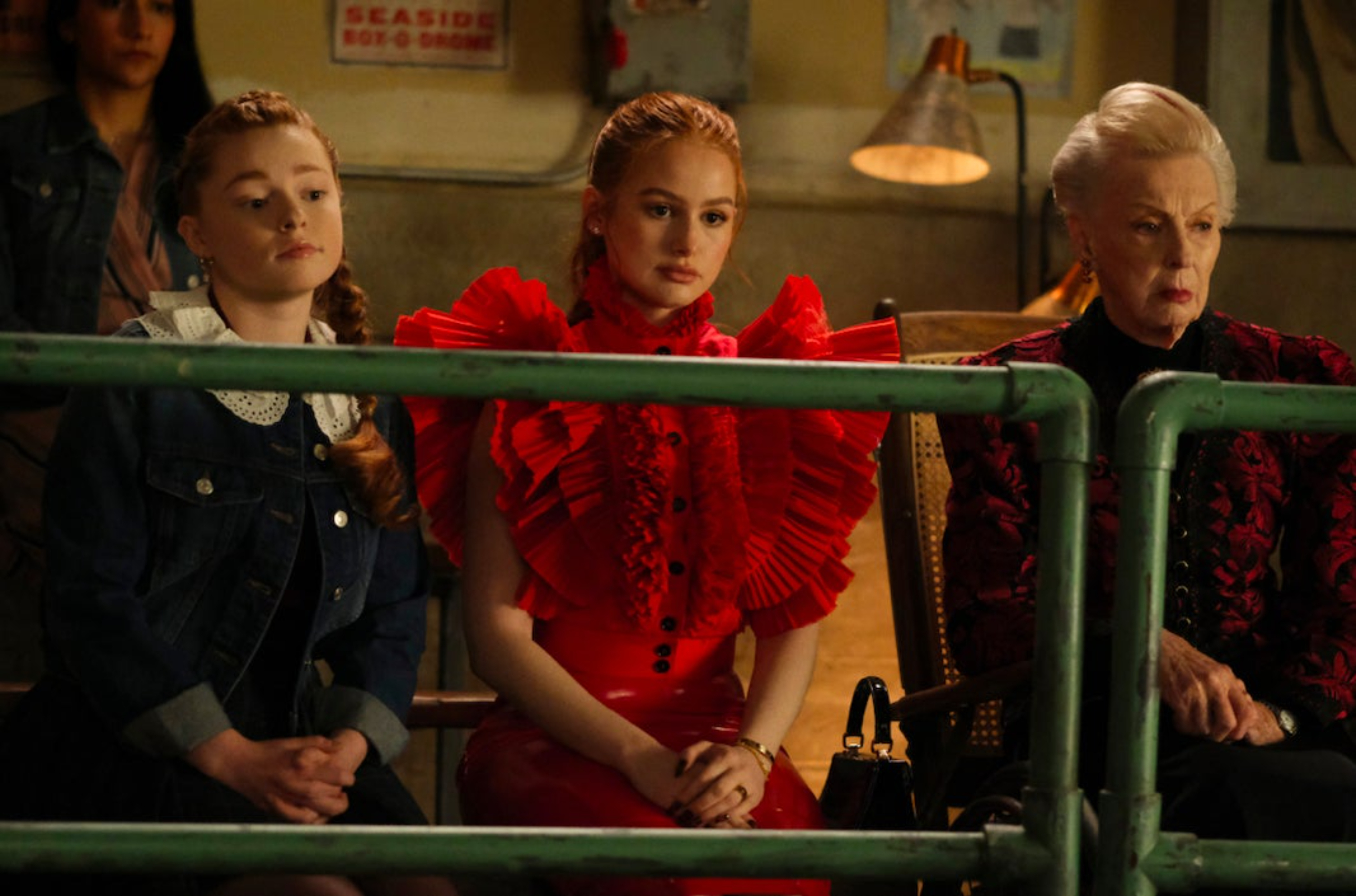 Three women sit behind a green metal fence. On the left is a redhead teenage girl, wearing blue denim. In the middle is a young redhead woman, wearing a red outfit. On the right is an elderly woman in dark red and black clothes.