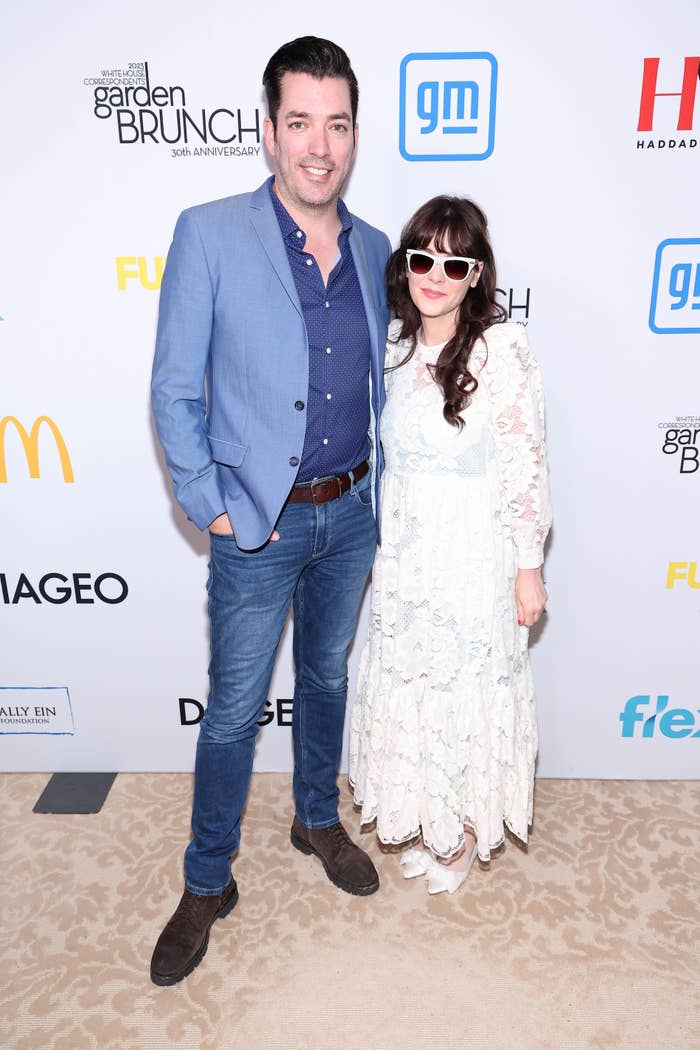 Jonathan Scott and Zooey Deschanel smile for photographers on the red carpet. Jonathan is wearing a blazer and jeans. Zooey is wearing a long lace dress, matching heels, and sunglasses