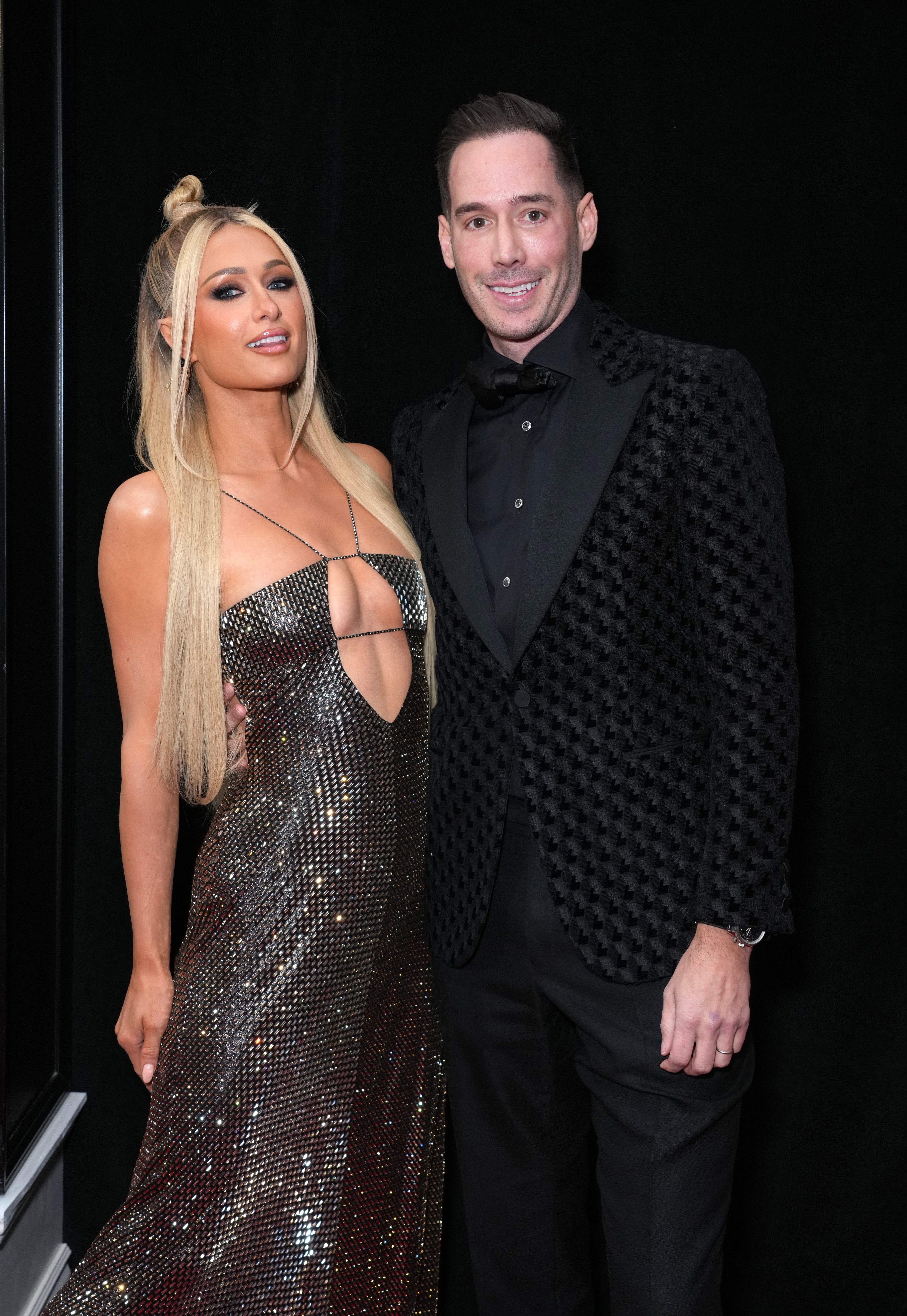 Paris Hilton and her husband Carter Reum smile for the camera at an event. Paris is wearing a long sparkly dress and Carter is in a suit sans tie