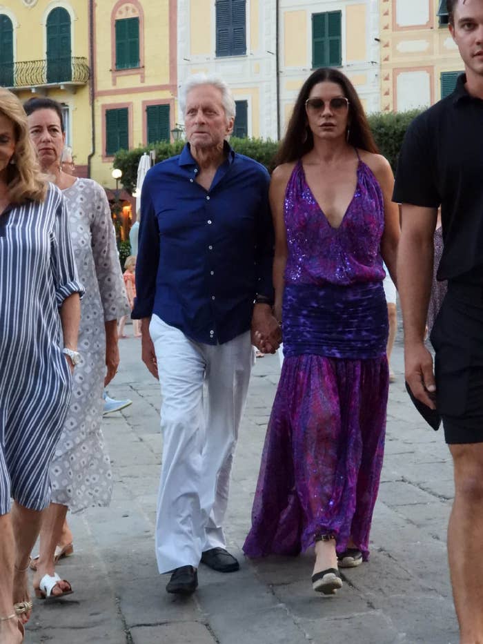 Michael Douglas and Catherine Zeta-Jones walking outside with several other people. Catherine is wearing a flow-y ankle-length dress with a dropped wait