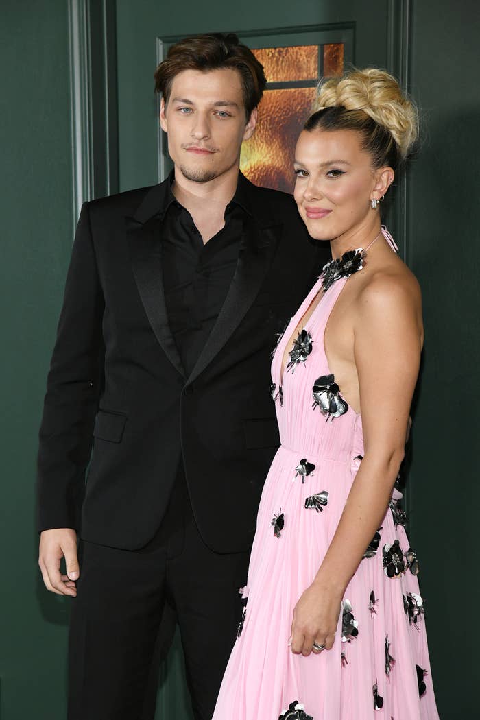 Millie Bobby Brown and Jake Bongiovi stand together for a photo at a media event. Millie is wearing an embellished halter dress and Jake is wearing a suit