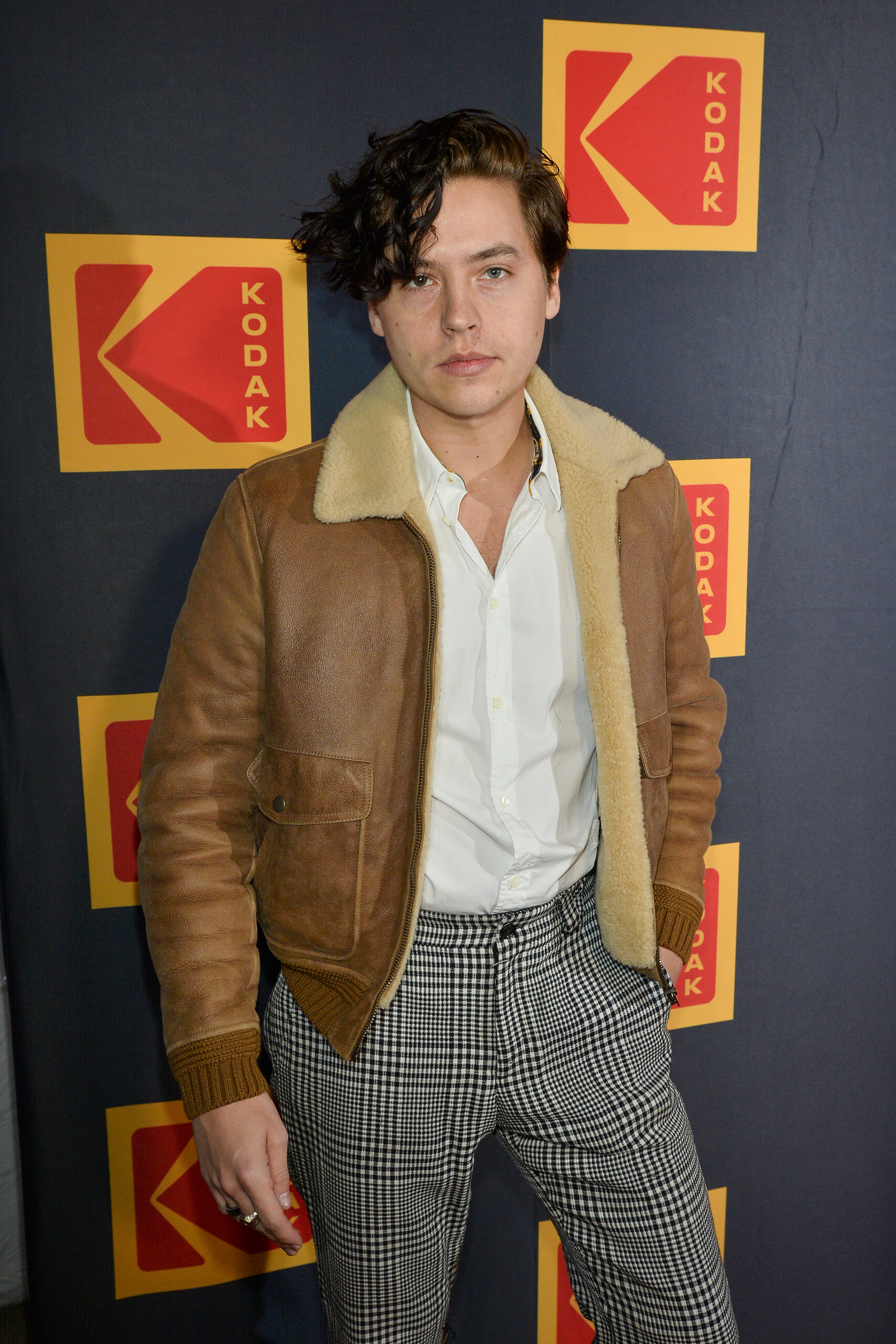 Closeup of Cole Sprouse at a media event wearing a shearling coat and slacks