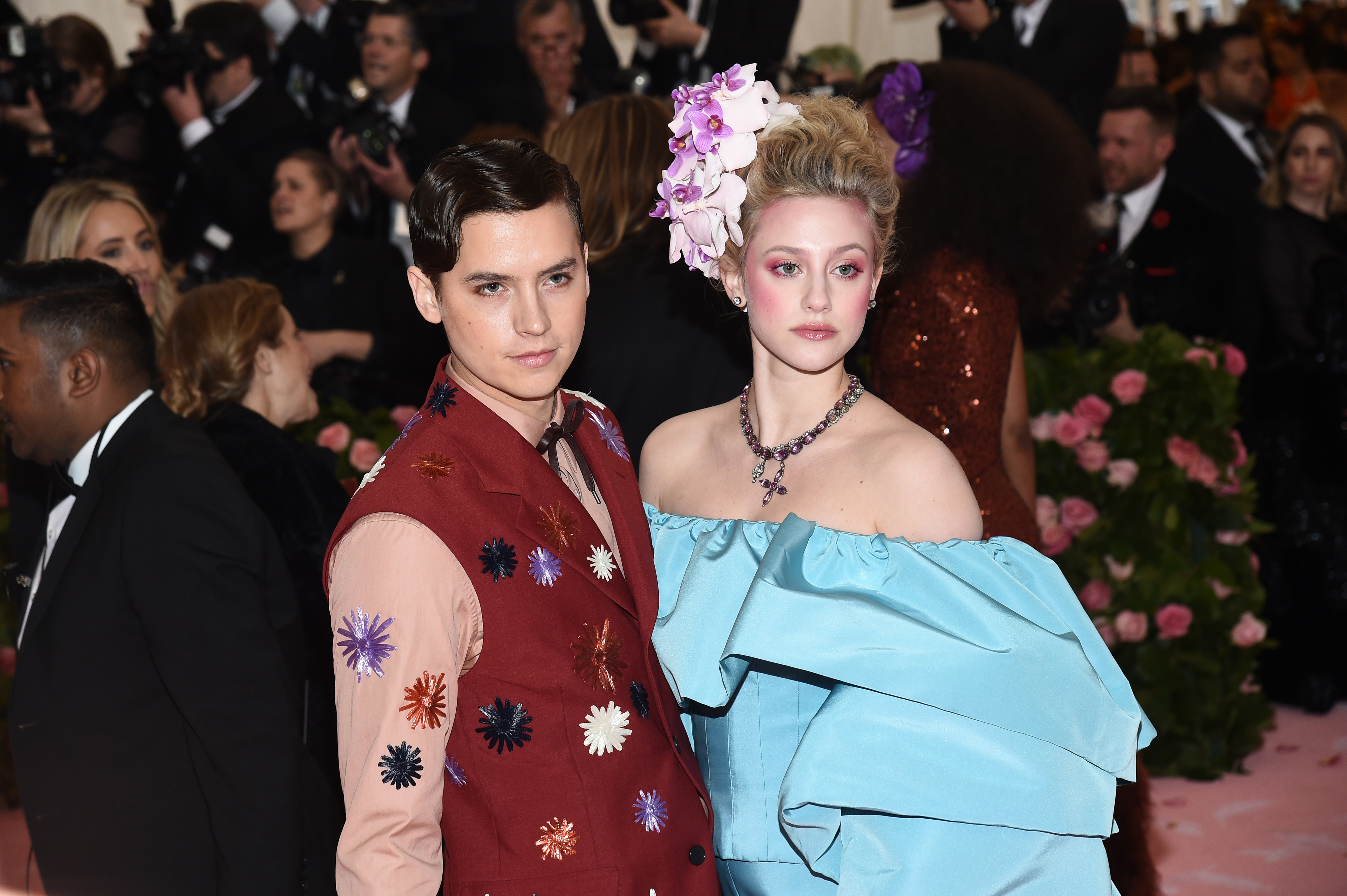 Cole Sprouse and Lili Reinhart on the red carpet. Cole is wearing a two-toned jacket with embroidered star bursts and Lili is wearing a off-the-shoulder dress with multiple flowers in her hair