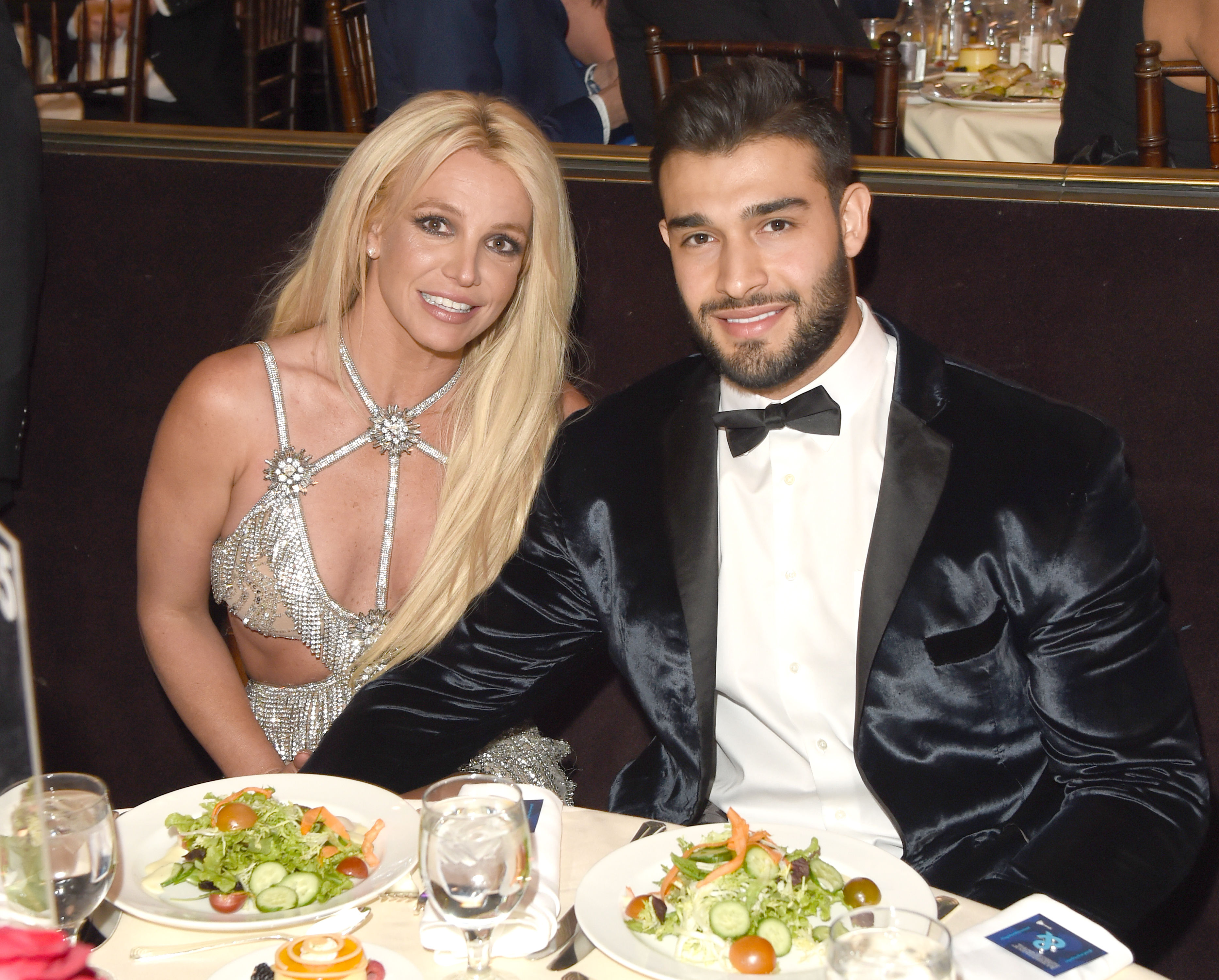 Britney Spears and Sam Asghari sitting at a table during an event with salads in front of them. Britney is wearing a sparkly with cutouts and Sam is wearing a tuxedo