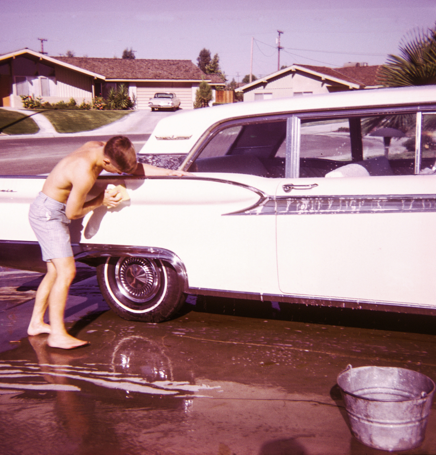 A teen is washing a big, &#x27;60s sedan in the driveway