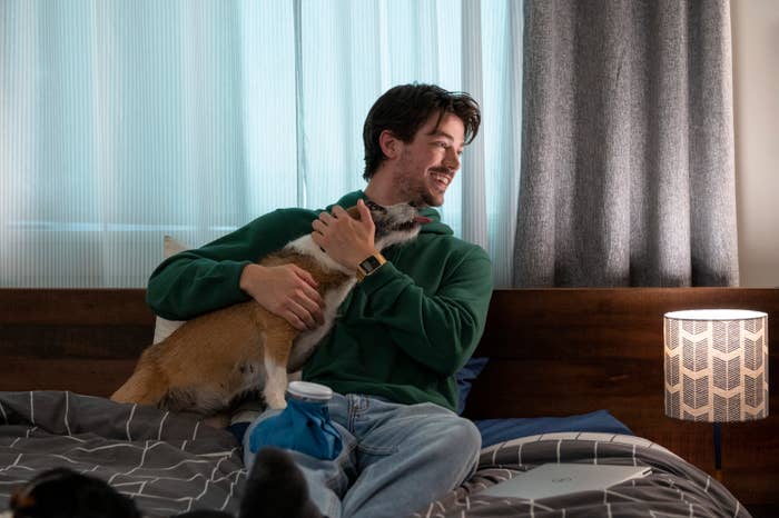 Grant Gustin as Max sitting on a bed with a hot water bottle on his knee as he holds a dog