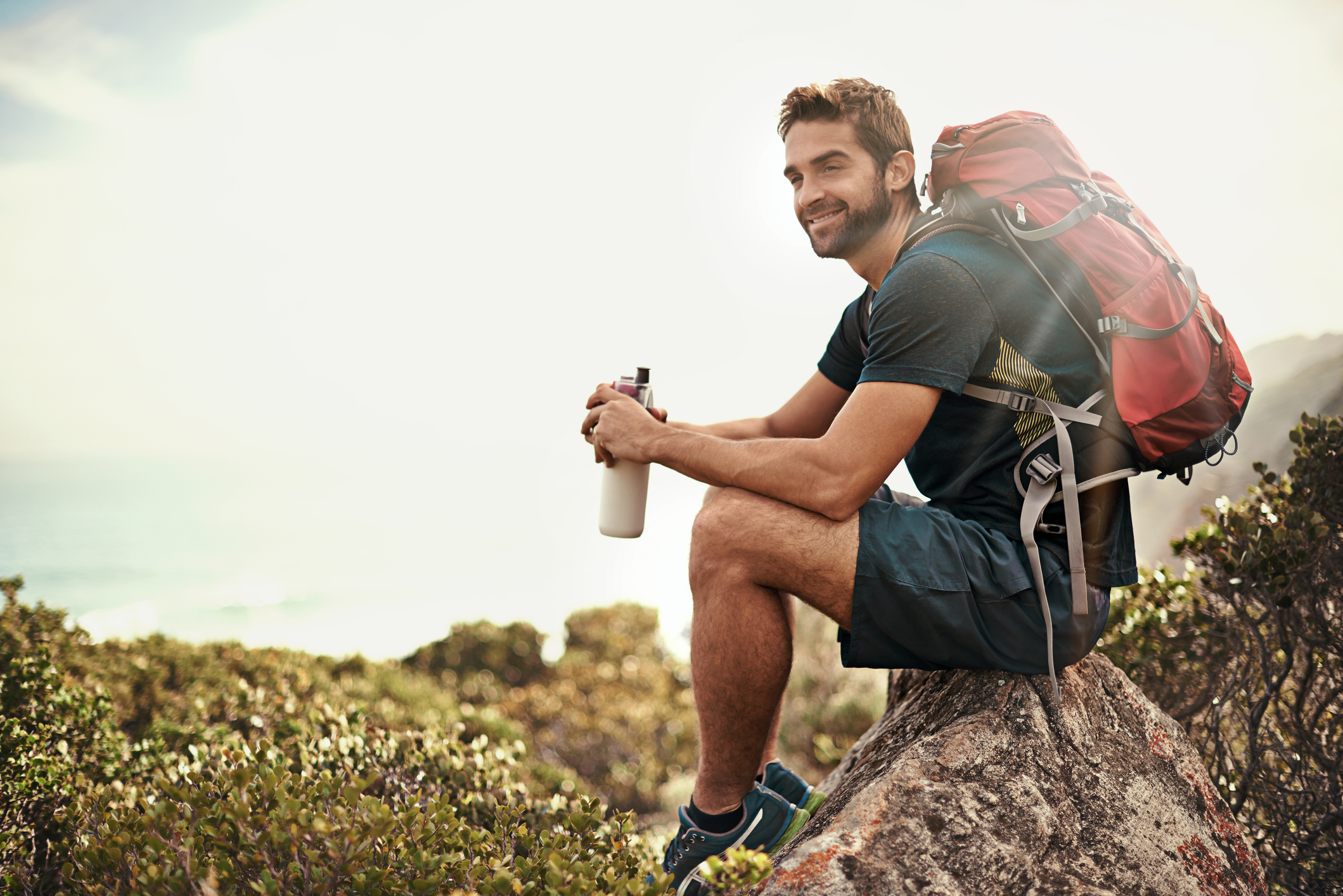 A smiling man in shorts with a backpack holds a water bottle and sits on a rock