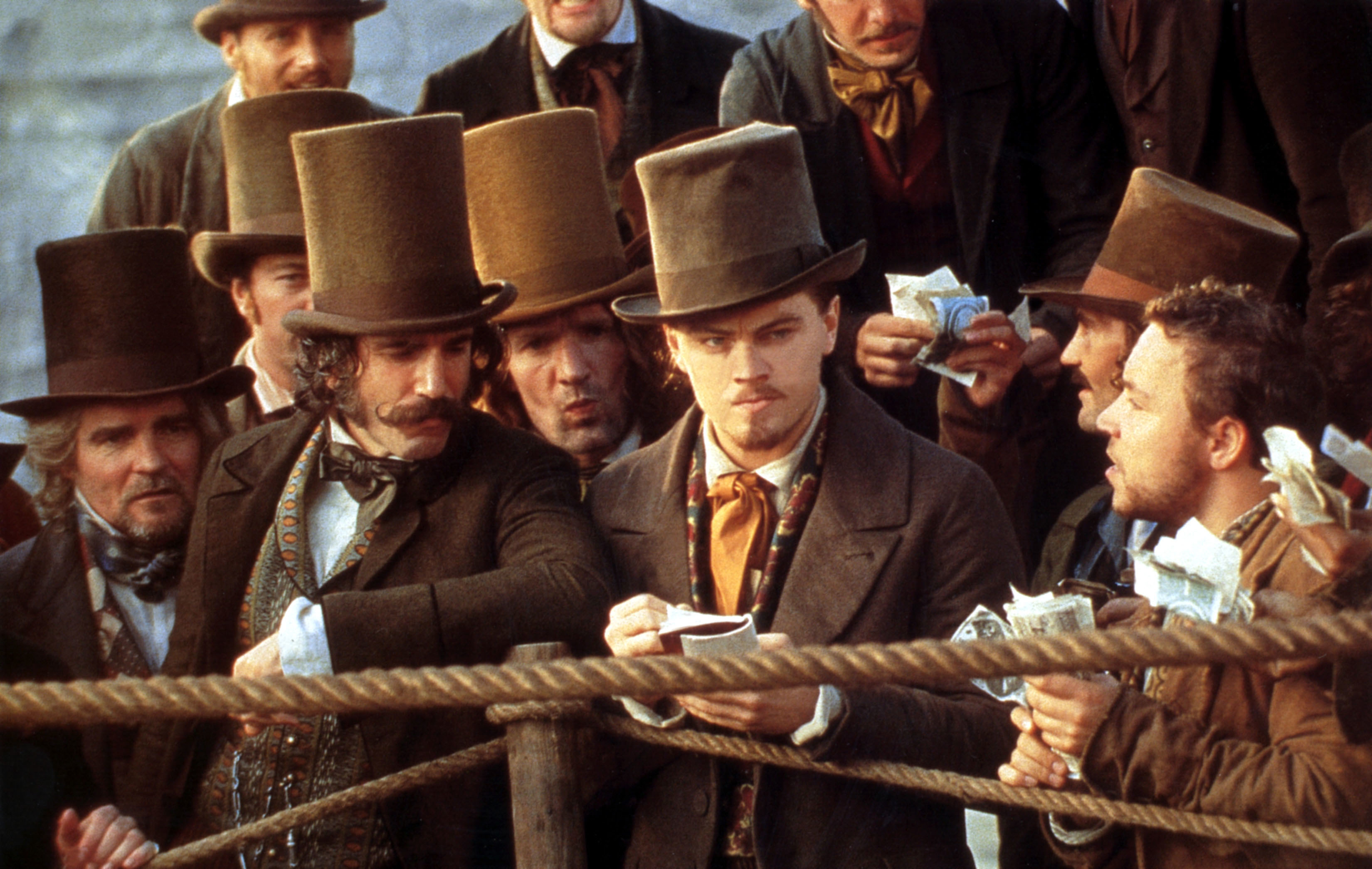 Daniel Day Lewis and Leonardo DiCaprio, donning top hats and brown jackets, stand near a rope barrier