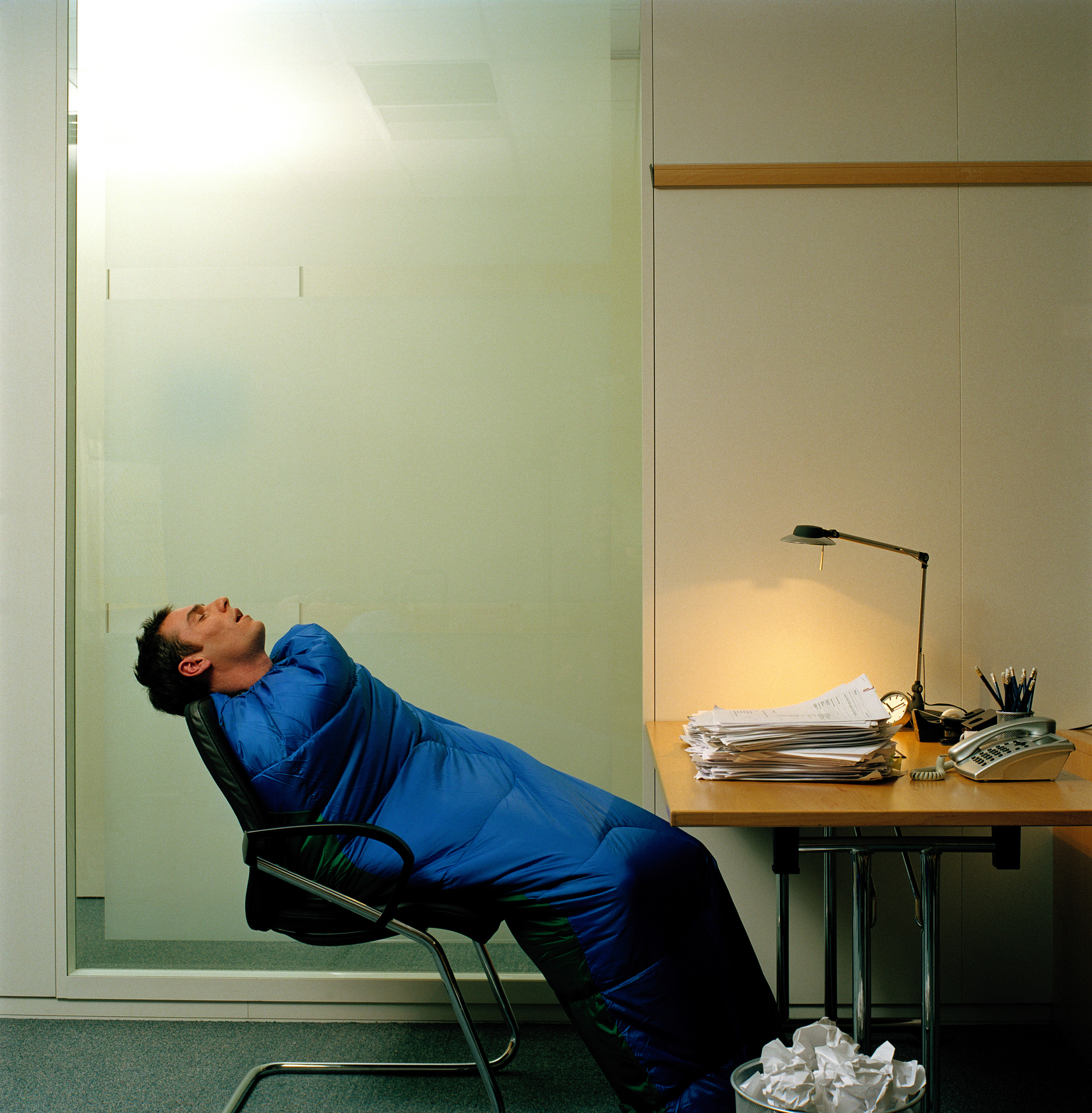 A man sleeping in an office chair while wrapped in a sleeping bag