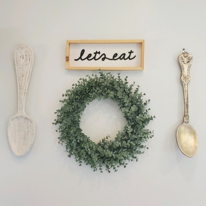The wreath hanging under a sign that says &quot;let&#x27;s eat&quot; and two spoon decorations on either side