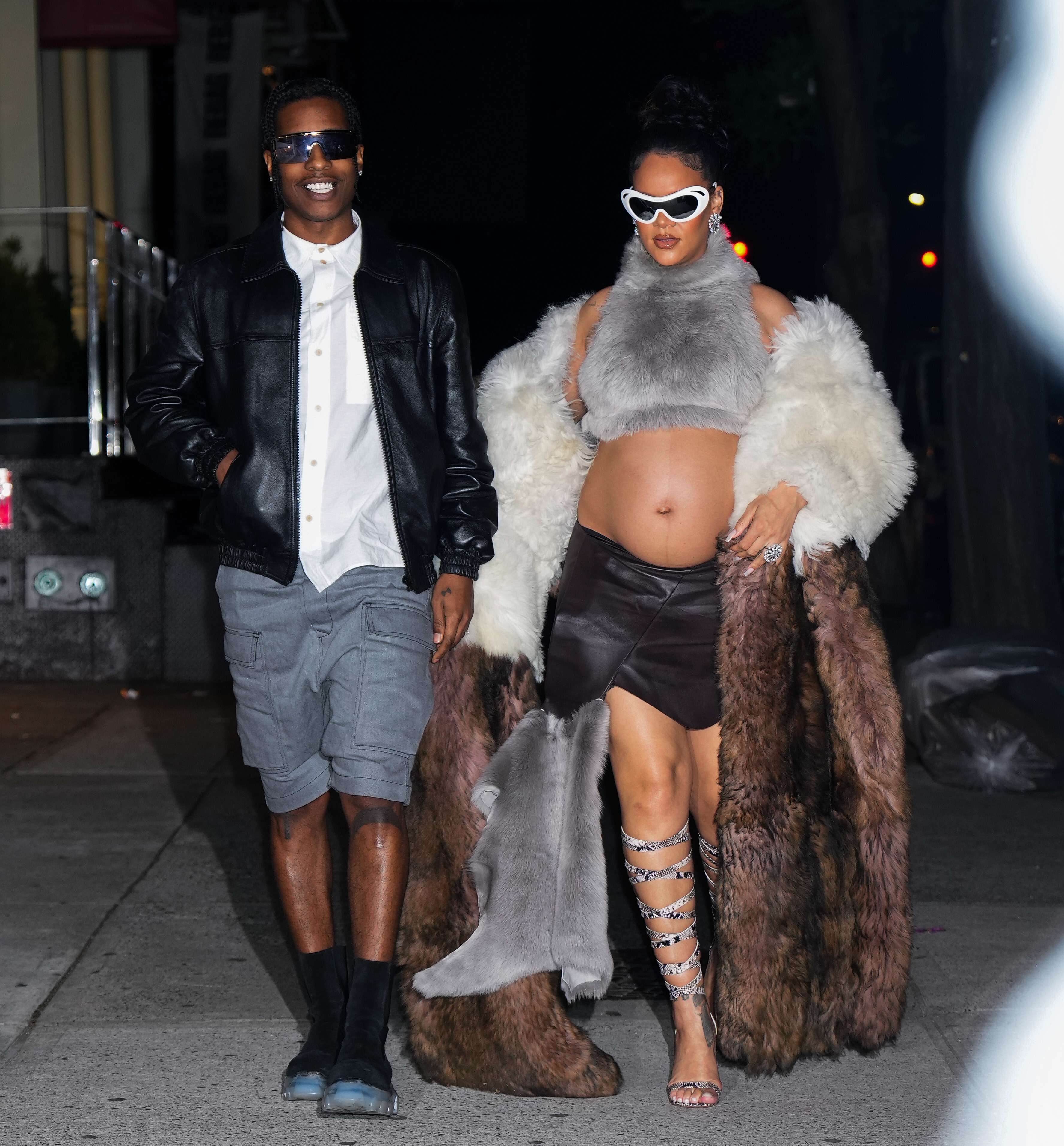 A$AP Rocky and Rihanna walking down the sidewalk at night. Rihanna is wearing a fur crop top and leather skirt with a fur coat. Rocky is wearing a dress shirt, shirts, and a jacket