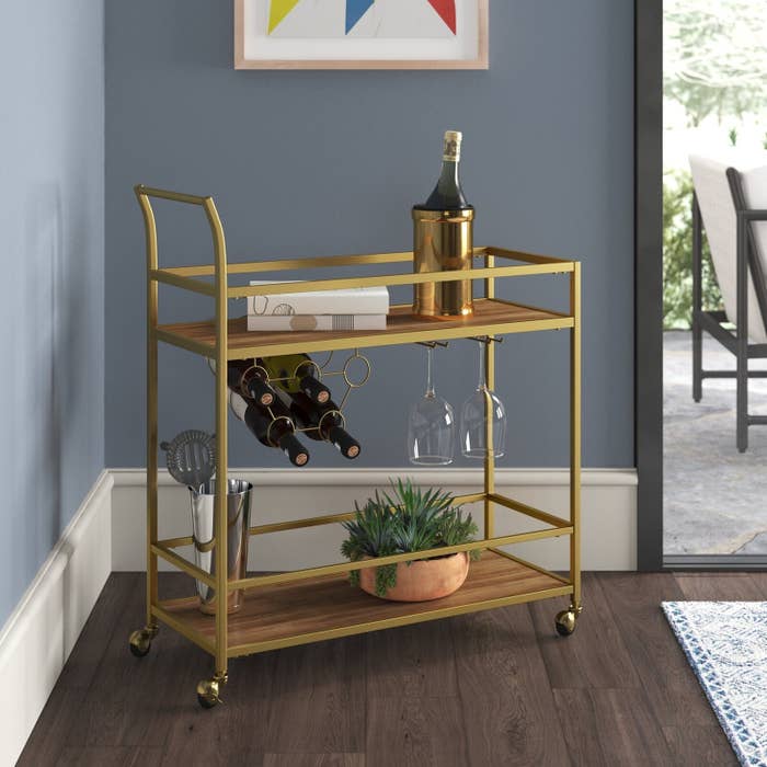 A gold bar cart with wine, champagne, and a plant in it.