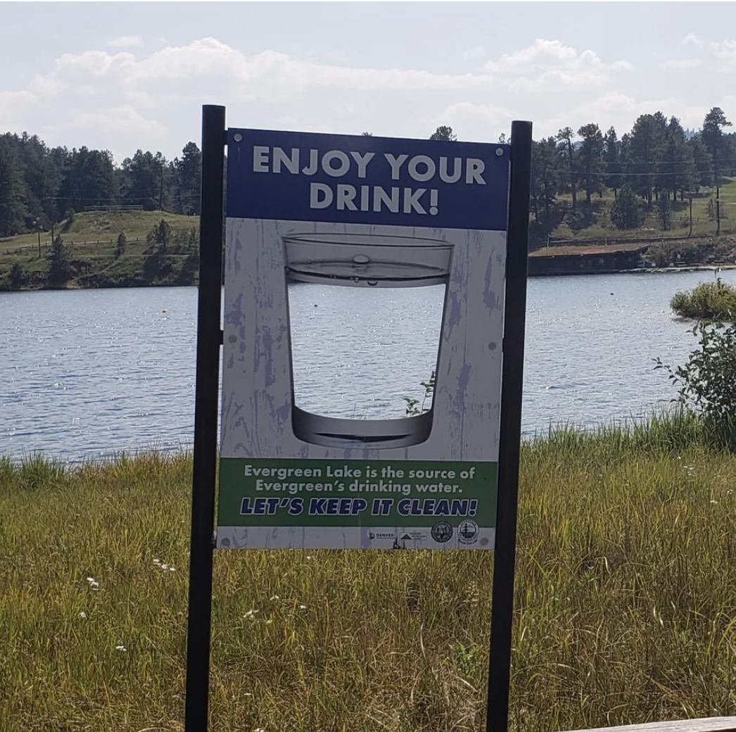 evergreen lake is the source of drinking water, let&#x27;s keep it clean, and the sign has a cut out of a cup with the lake in the back filling it in