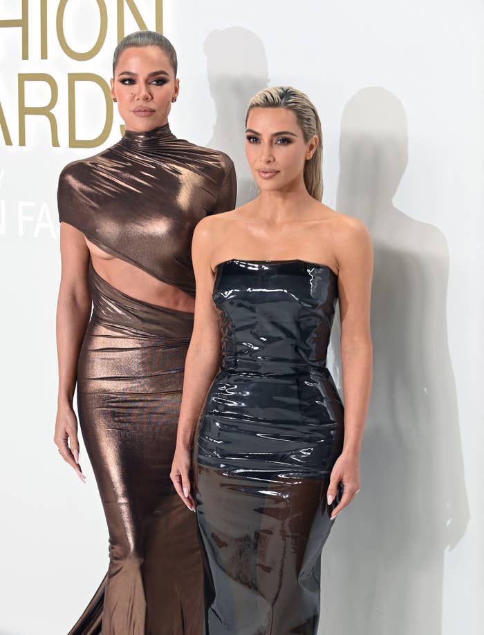 Khloé and Kim Kardashian on the red carpet. Khloé is wearing a metallic dress with a cuotout across the abdomen and Kim in wearing a pleather strapless dress