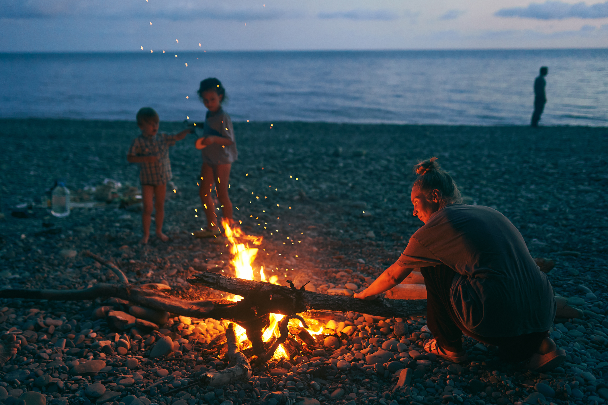 A woman by campfire with two children at the beach at night