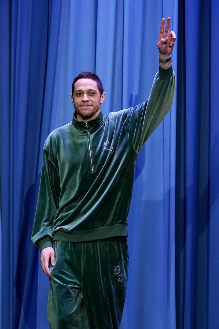 Pete in a velour tracksuit raises his hand in greeting on a talk show