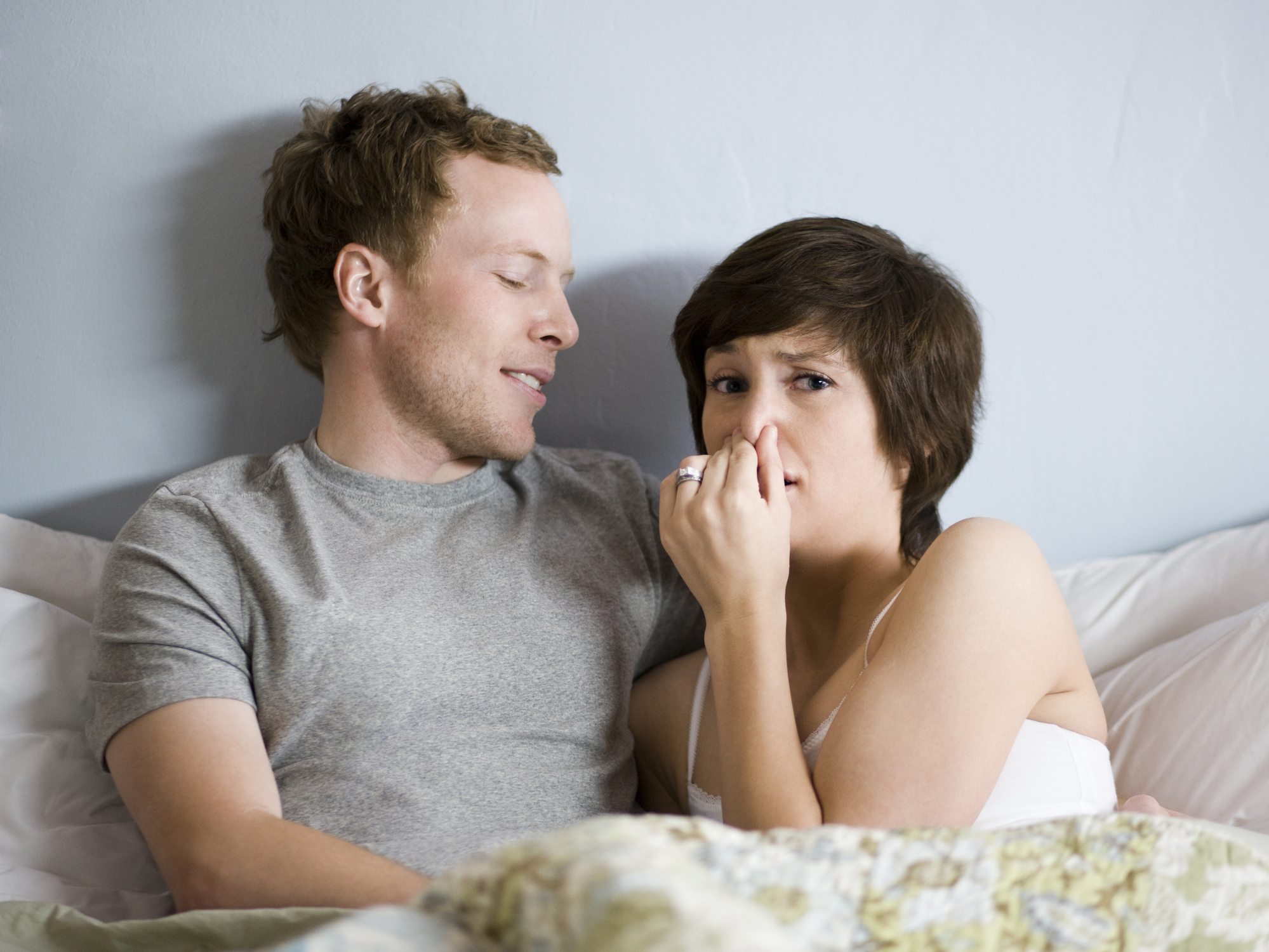 A man and a woman sitting up in bed and the woman has her fingers covering her nose