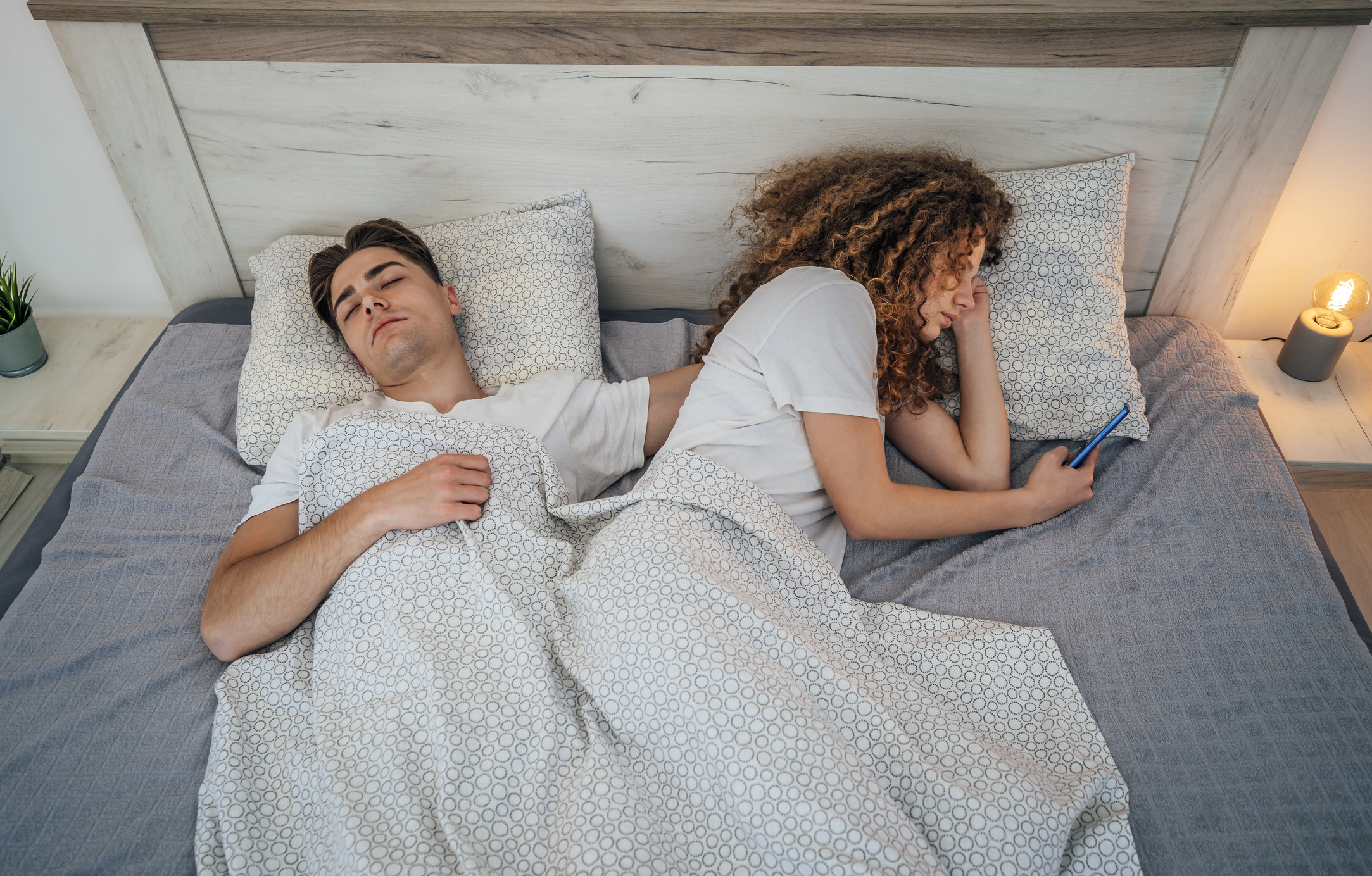 A woman facing away from a man in bed and looking at her phone