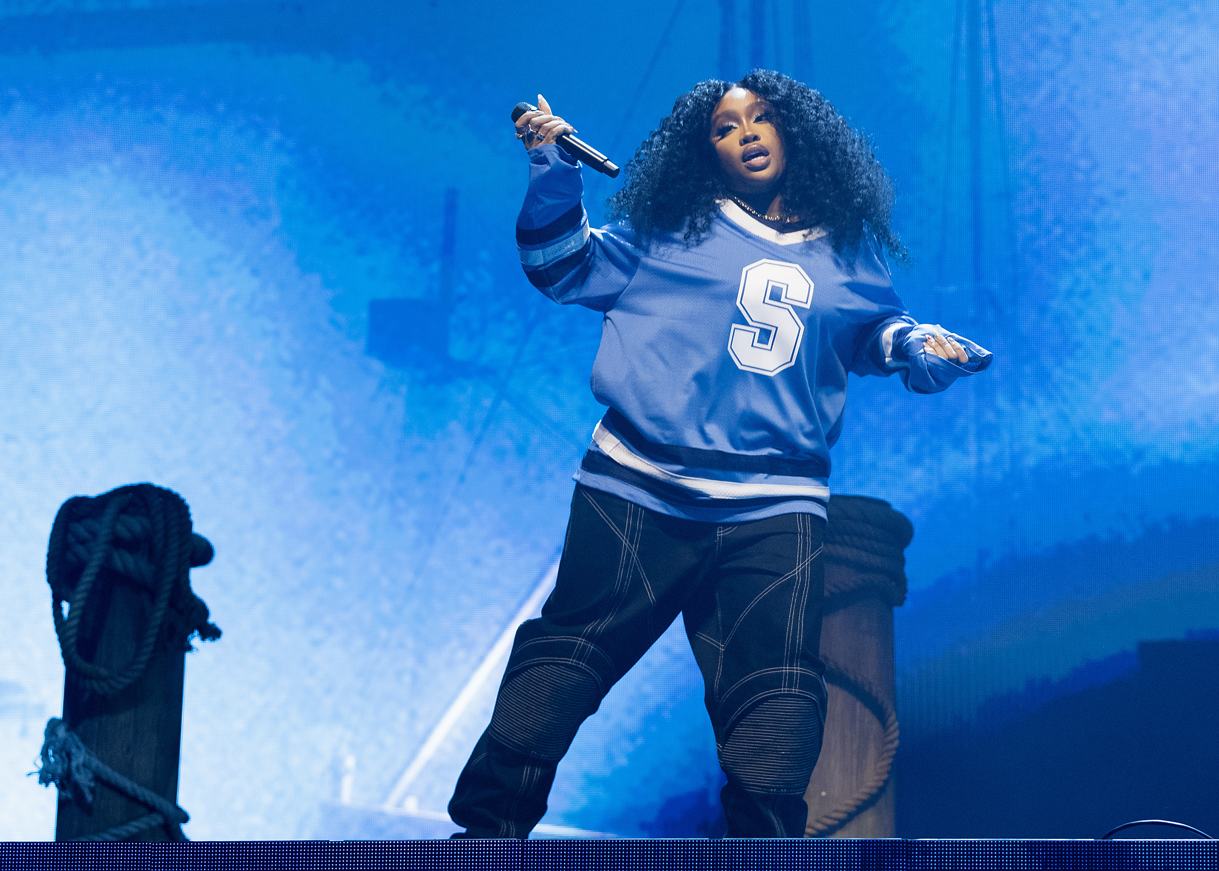 SZA performing onstage in loose jeans and a jersey