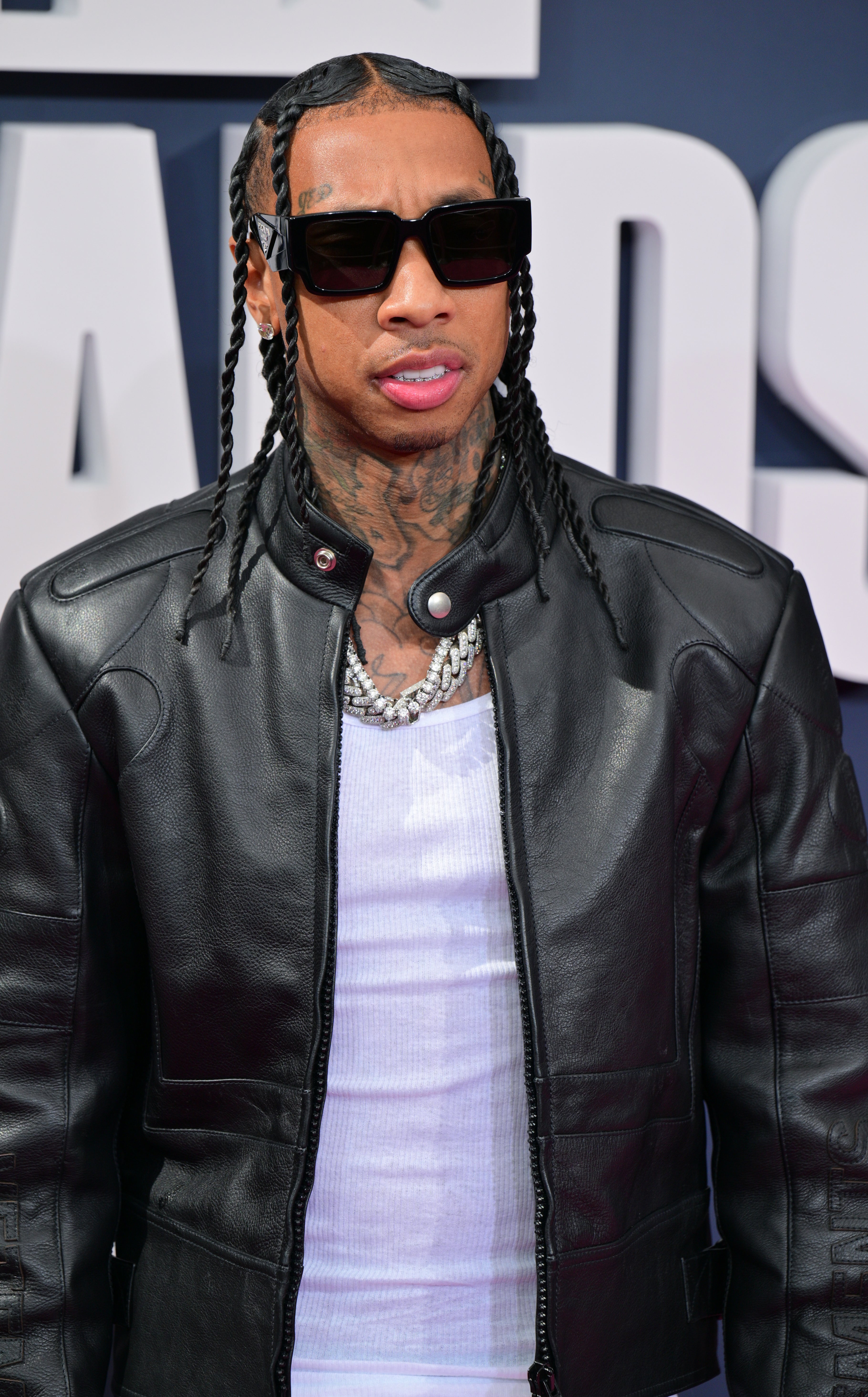 Tyga on the red carpet wearing a shirt, chain, leather jacket, and sunglasses