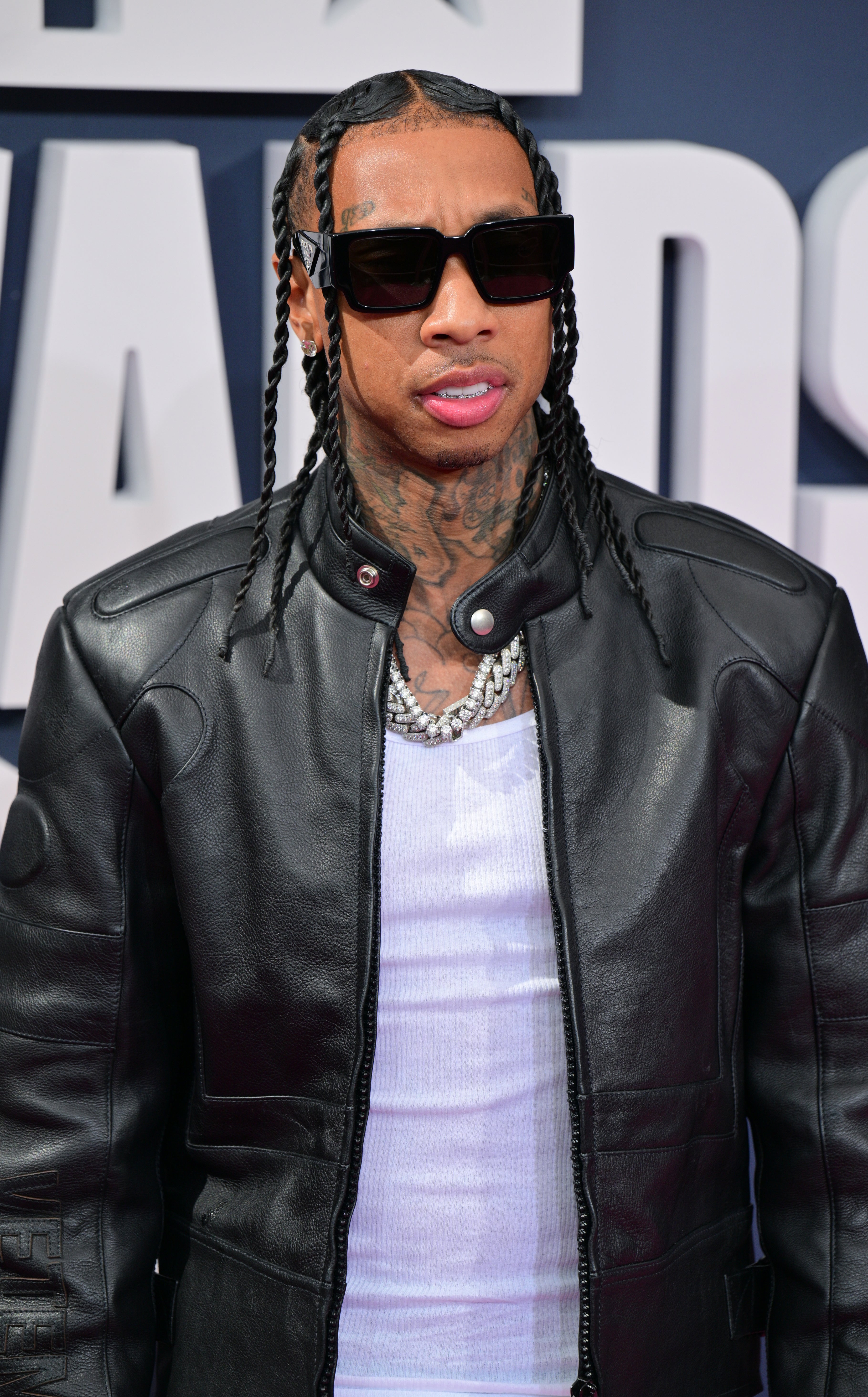 Tyga on the red carpet wearing a shirt, chain, leather jacket, and sunglasses