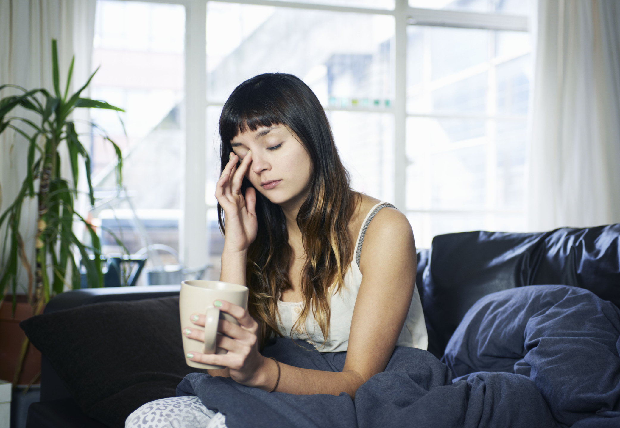 A tired looking woman sits on her couch and rubs her face while holding a mug