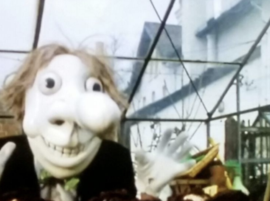 A paper white puppet with a large bulge attached to his nose, a large, creepy smile, bulging eyes, and very unkempt hair