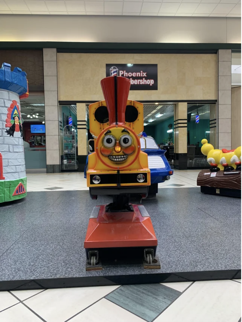 A small children&#x27;s stationary train ride in a local mall that has big, wide eyes, defined rosy cheeks, and a huge, scary smile