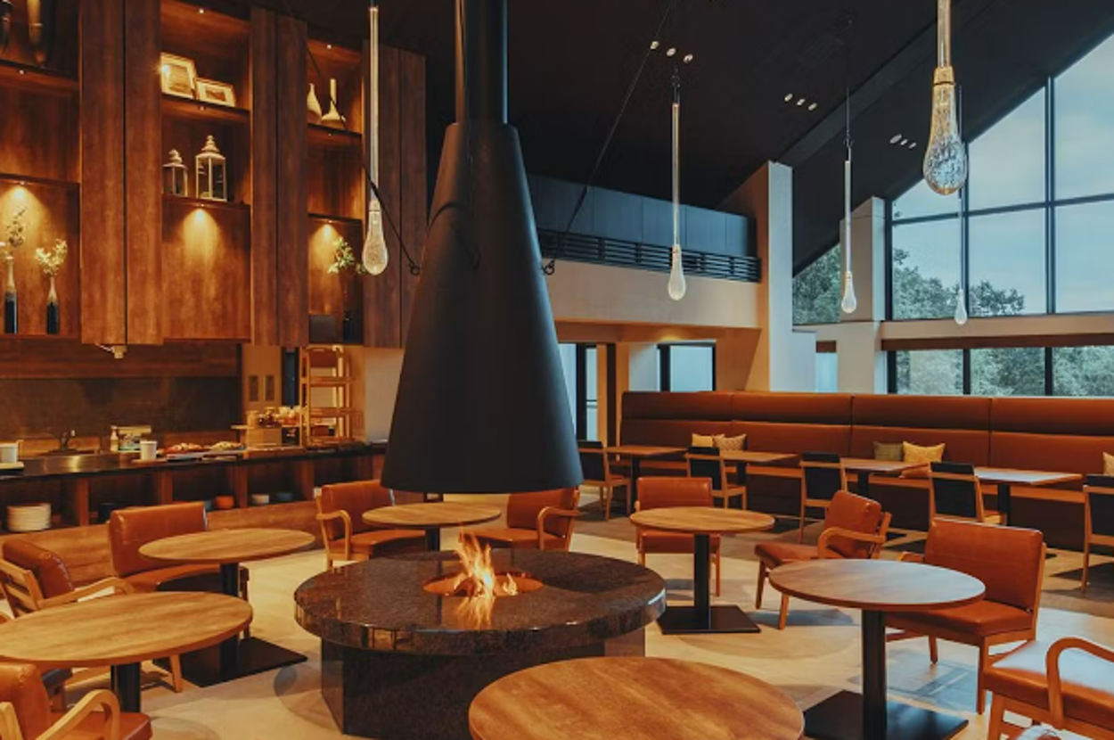 高級感のあるカフェラウンジ、暖炉を囲む座席とモダンな照明。広い窓からの自然光が心地良い雰囲気を演出。