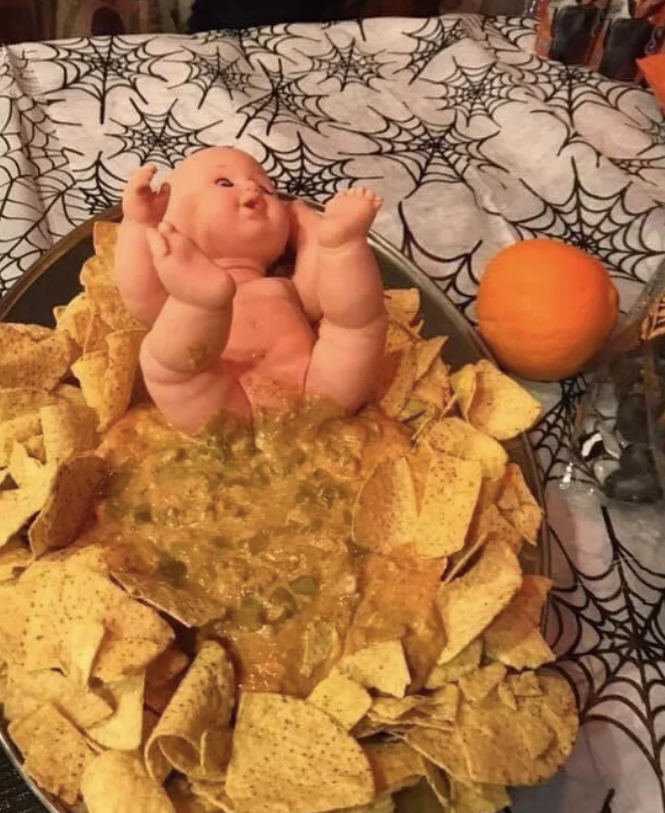 A naked baby doll on its back with its legs up in the air at the top of a large bowl, with liquidy, yellow-green dip extending from its bottom and surrounded by chips