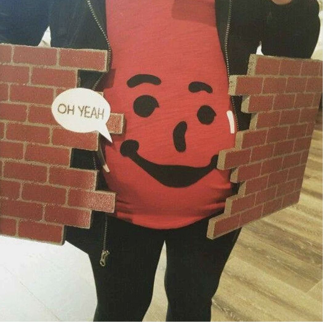 Pregnant person wearing a Kool-Aid man&#x27;s face on a shirt for Halloween with &quot;Oh yeah&quot; dialogue bubble emerging from the mouth, along with pieces of a brick wall on either side of the stomach