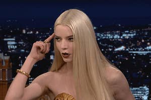 Anya Taylor-Joy holds her pointer finger to her head and leans forward.