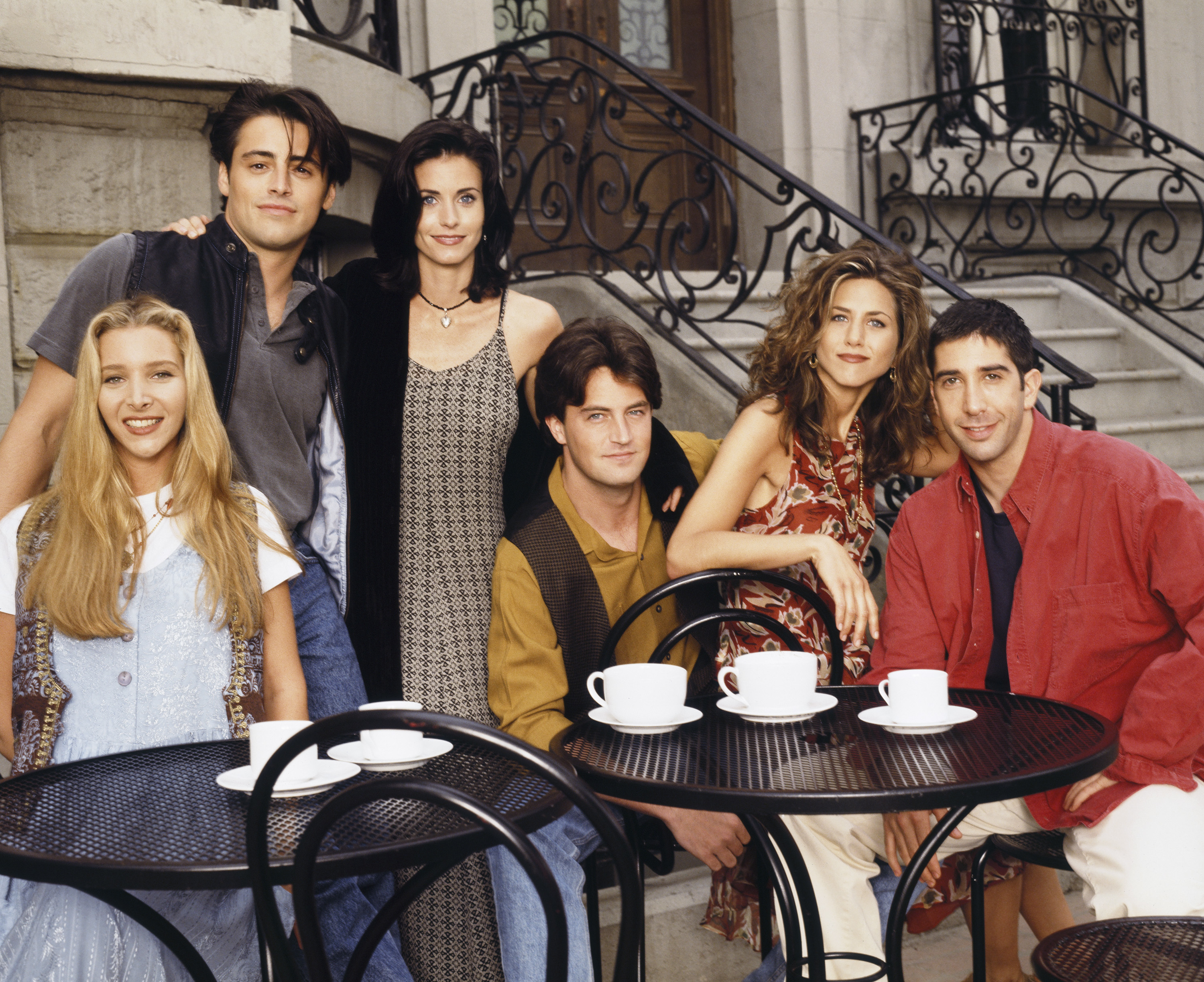 The six leads of Friends sitting or standing together at several tables